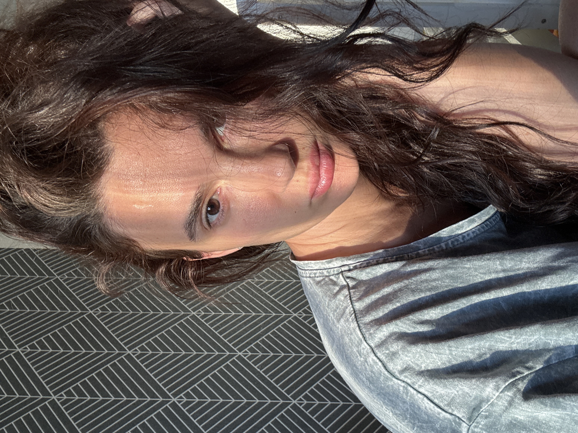 The image shows a close-up, medium shot of a young adult with long, dark, wavy hair. The person is looking directly at the camera with a neutral expression. Sunlight is casting a strong shadow across their face, highlighting their cheekbone and nose. They are wearing a light blue t-shirt. The background features a dark gray wallpaper with a geometric pattern of white lines that resemble stylized leaves or feathers. The lighting suggests the sun is positioned to the right of the subject, creating a warm, golden hour glow on their skin. There are no discernible landmarks indicating a specific location within the city of Chaikovsky, Russia.