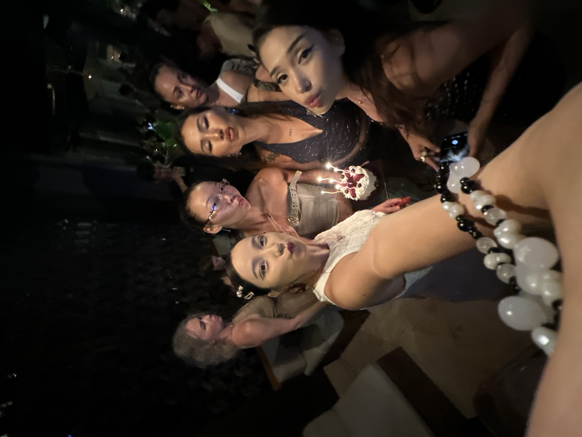 A group of six women is taking a selfie with a small birthday cake with lit candles. The photo appears to be taken in a dimly lit outdoor setting, possibly a restaurant or bar. The location is inferred to be North Kuta, Indonesia. The woman in the foreground is holding the camera and has a beaded bracelet on her wrist. The cake is being held by one of the women in the middle of the group. Some of the women are looking at the camera. There are dark areas indicating nighttime. The scene appears to capture a birthday celebration among friends.