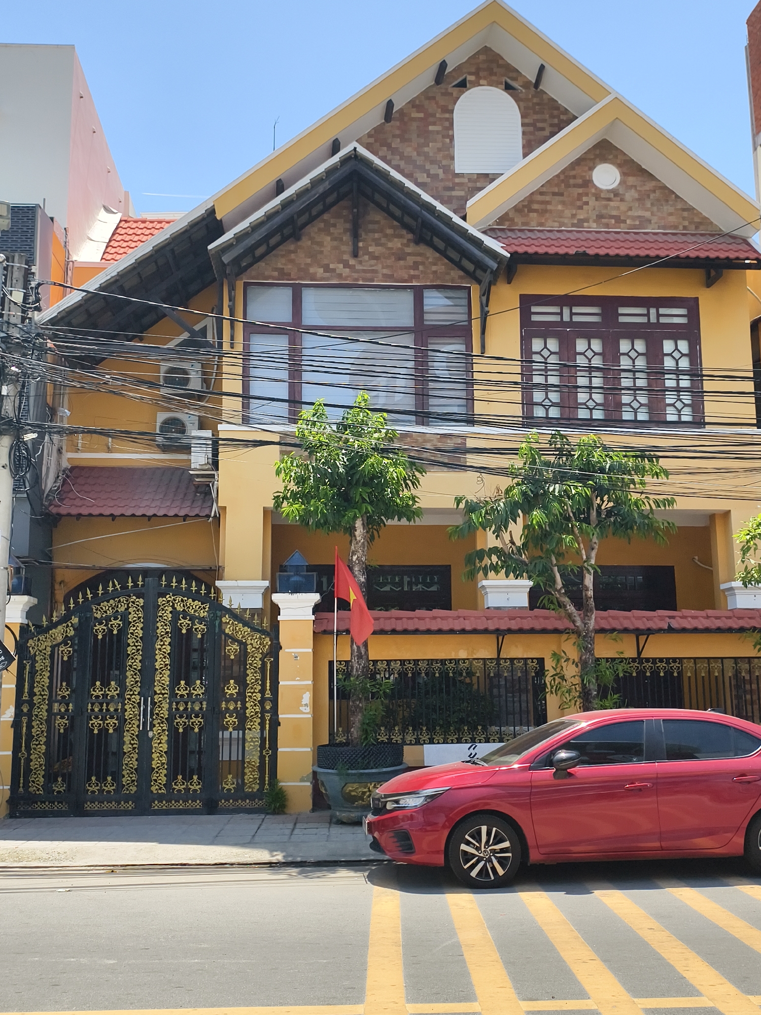 The scene depicts a yellow multi-story house situated in Tay Nha Trang Ward, Vietnam, under a clear blue sky. The building features a prominent facade with a gabled roofline, dark red roof tiles, and sections of exposed brick-patterned walls on the upper gables. White trim outlines the roof edges and windows.
On the upper level, there are two distinct window arrangements: a large, multi-pane window with dark frames on the left and a wider window with decorative grid patterns and dark frames on the right. Above these, the gables contain a white arched vent on the left and a small circular vent on the right. Below the main roof, smaller tiled awnings extend over the first floor.
The ground level features a large, ornate black metal gate with intricate gold accents, opening onto a concrete sidewalk. To the right of the gate, a smaller black metal fence extends along the property line. A Vietnamese flag, red with a yellow star, is displayed on a pole near the fence. Several small trees with green foliage are planted in front of the house, some in decorative pots.
Numerous black utility cables crisscross overhead, particularly dense on the left side of the image, where multiple outdoor air conditioning units are mounted on the house's exterior. To the far left, a portion of an adjacent building with a light-colored facade is visible.
A red sedan, identifiable as a Honda City, is parked on the asphalt street in the foreground, partially overlapping a yellow-striped pedestrian crossing. The car faces left, with its side profile visible. The sky above is clear, with a faint contrail from a distant aircraft visible. The overall lighting suggests a bright, sunny day.