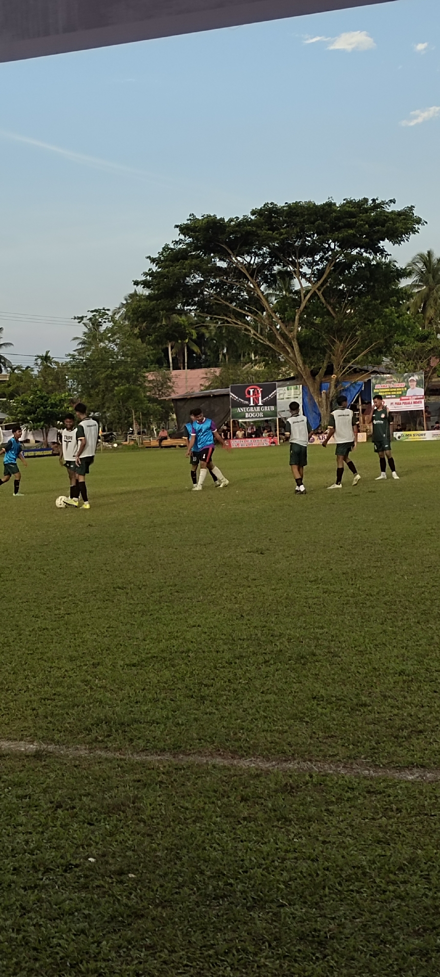 The image depicts a soccer game on a grass field. Several young men in athletic uniforms are on the field, some with a soccer ball. In the background, there are trees, buildings, and banners. One banner reads "ANUGRAH GRUB BOGOR". The sky is partly cloudy. The overall scene suggests an outdoor sporting event in a semi-urban or rural setting, likely in Kilometer Delapan, Indonesia, given the banner.
