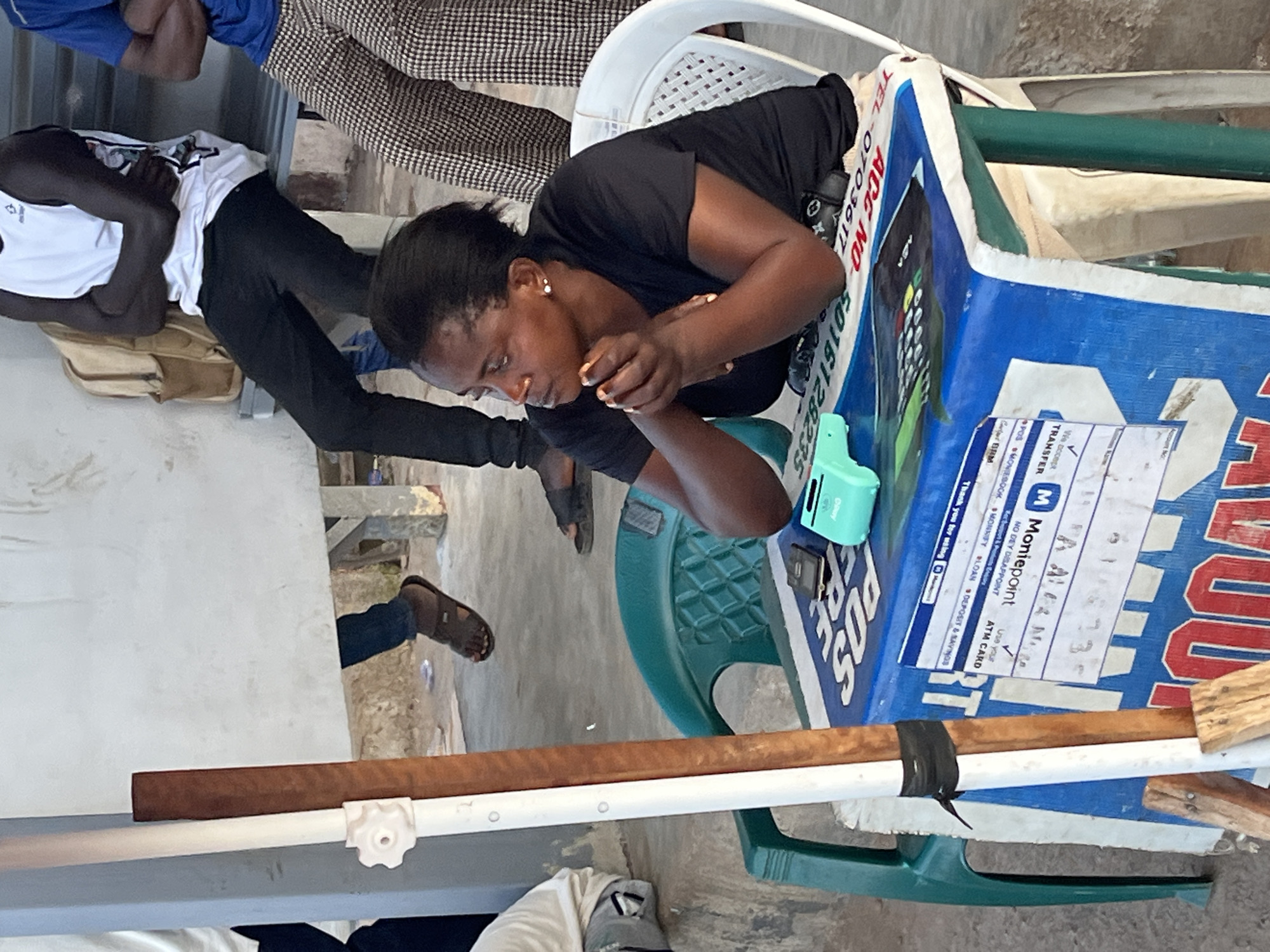 This media file depicts a scene with several individuals in an outdoor setting, likely in Gura Top, Nigeria. The primary subject is a woman with dark hair, wearing a black t-shirt, who is leaning over a blue table. Her hands are engaged, and she appears to be eating or holding something small. Her expression suggests focus or concentration.
Beside her, a person wearing a white tank top and black pants is seated, with their arms crossed and their head resting on their arm. Another person, partially visible in the background, wears a patterned outfit and appears to be seated. The setting includes a light-colored table and a green plastic chair.
The blue table has various pieces of paper and a small teal device on it. Visible text on the blue table includes "POS", "TRANSFER", "Moniepoint", "ATM CARD", "LOAN", and "DEPOSIT & SAVINGS". Numbers and text like "ACC NO 5016128235" and "TEL-0703617" are also legible. The overall atmosphere seems casual, possibly a marketplace or a public gathering space. The lighting suggests it might be daytime.