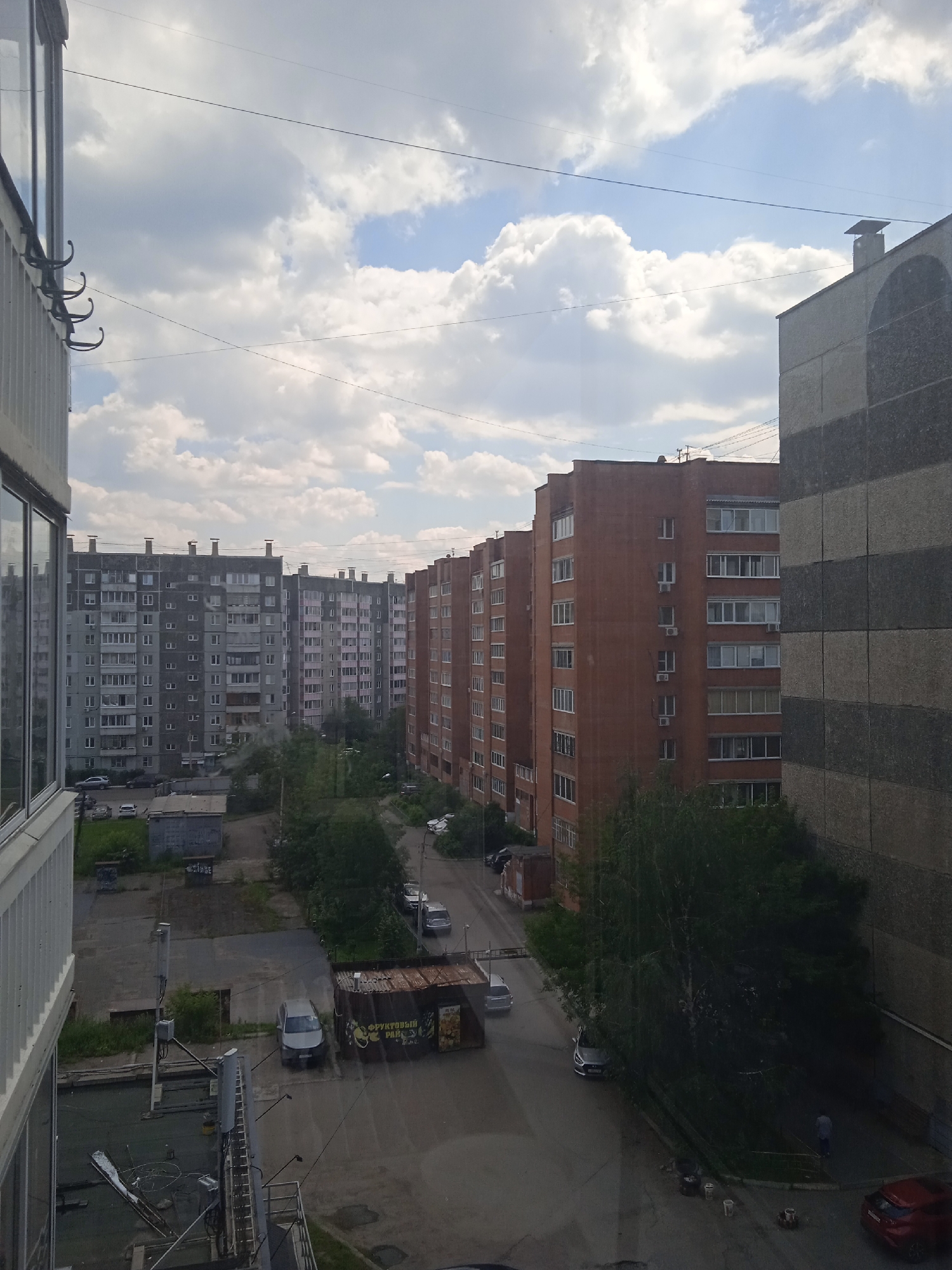 The image depicts a view from an upper-story window overlooking a residential area in Krasnoyarsk, Russia. Several multi-story apartment buildings are visible, constructed from grey and orange brick, and concrete panels. Some buildings have visible chimneys on their roofs. The sky is partly cloudy, with large white cumulus clouds against a blue background. Several power lines crisscross the sky. Below the buildings, there is a paved courtyard with parked cars, including a silver sedan and a red hatchback. A small, dilapidated kiosk with Cyrillic lettering, possibly a fruit stand, is situated in the courtyard. Trees and greenery are scattered throughout the area, with a dense cluster of trees partially obscuring a courtyard. A person in a light-colored shirt is walking on a paved area near the trees. The scene appears to be an ordinary urban residential environment under an overcast sky.