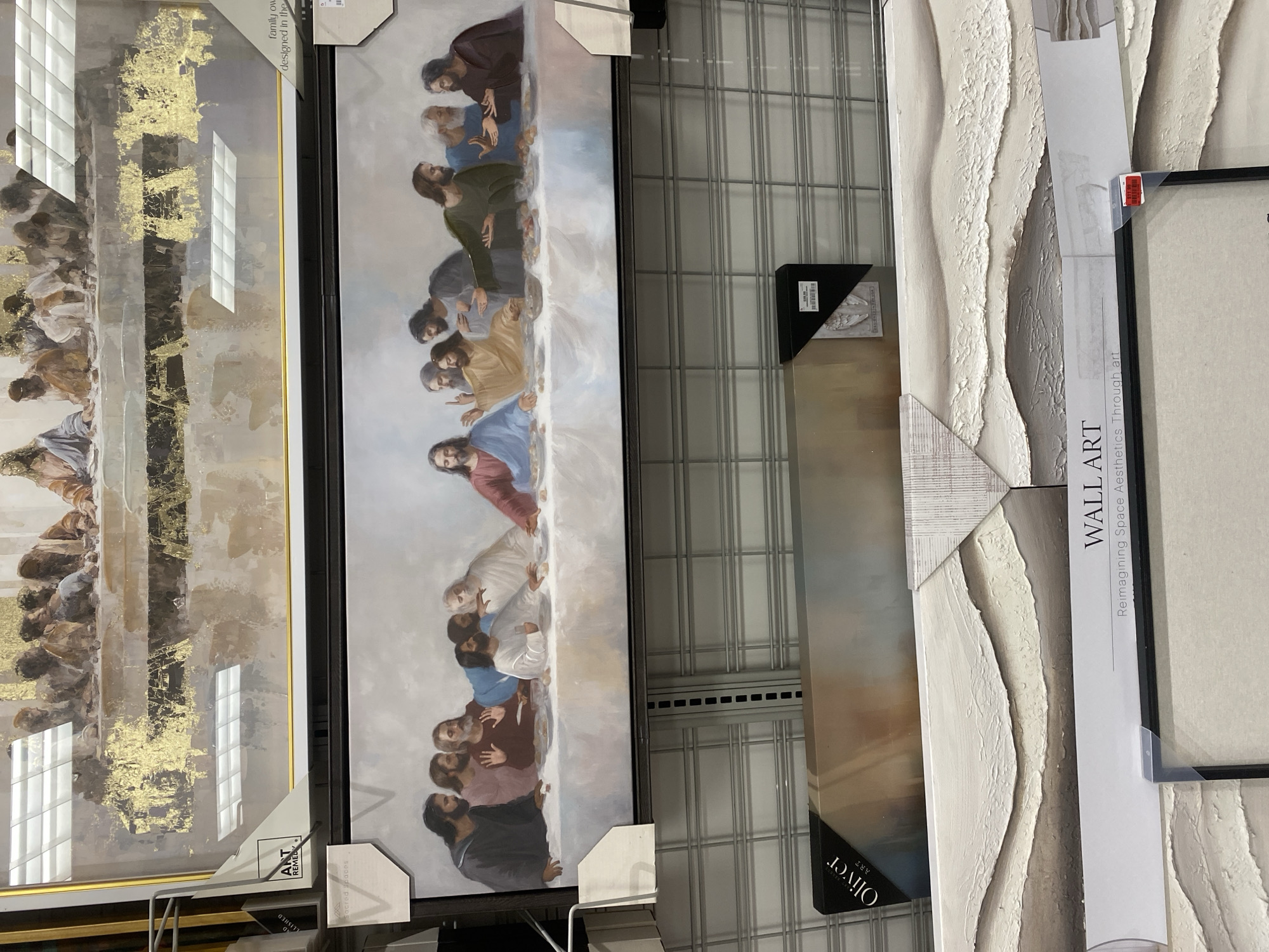 The image displays a retail store environment, likely a department store, showcasing various wall art pieces. In the foreground, a framed canvas print depicting "The Last Supper" by Leonardo da Vinci is prominently displayed. This artwork features Jesus and his disciples gathered around a table, rendered in a realistic style with a muted color palette. Above this print, another canvas print, seemingly an abstract depiction of "The Last Supper" with gold leaf accents, is visible. The background shows metal shelving units typically found in retail settings, with more artwork and boxed items stacked on them. A sign that reads "WALL ART Reimagining Space Aesthetics Through Art" is visible at the bottom. The presence of price tags and the overall display suggest the scene is inside a store in Inglewood, California, USA, where such merchandise is sold.