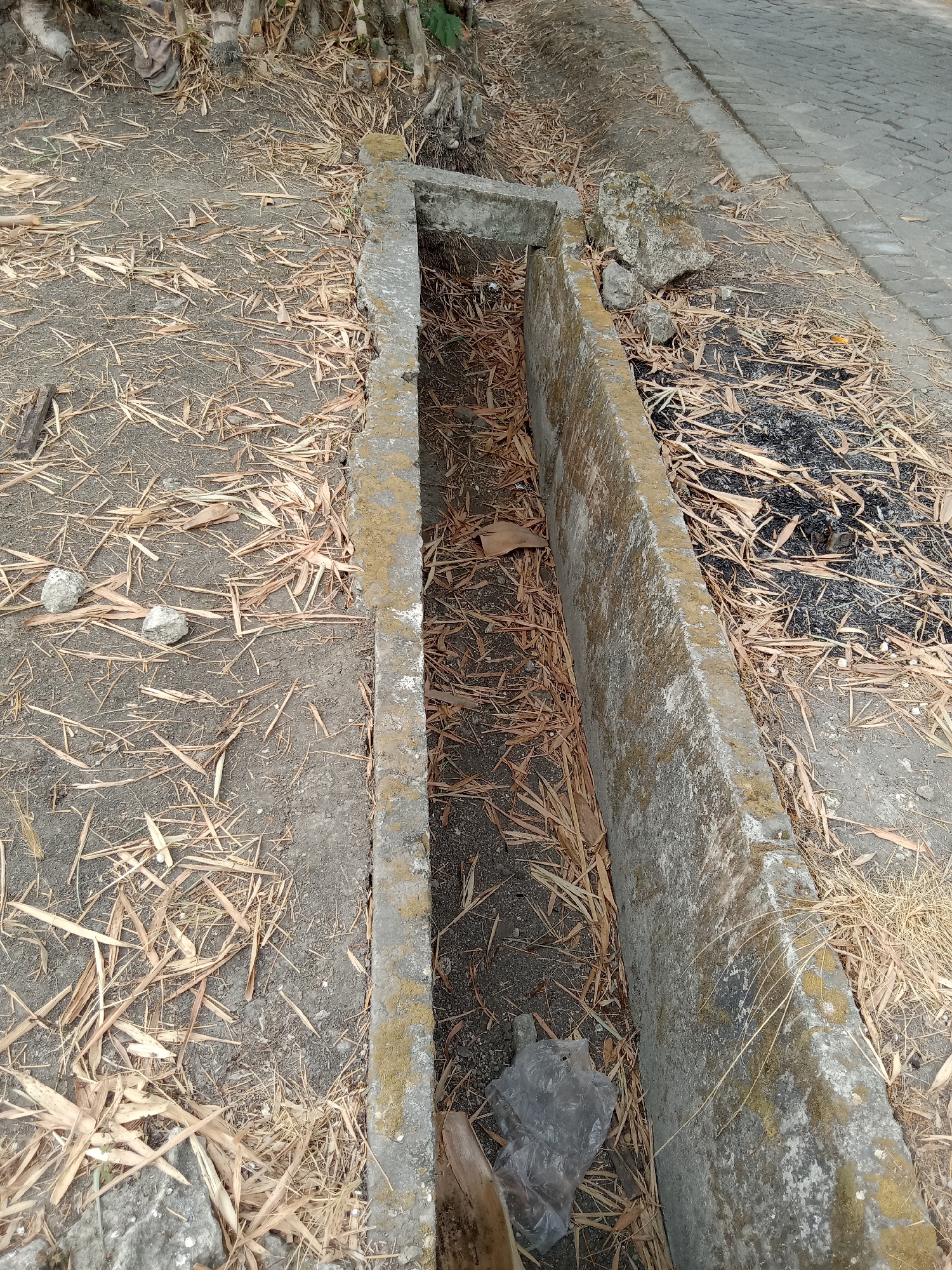 The image depicts a concrete drainage ditch filled with dry leaves, debris, and a clear plastic bag. The ditch runs alongside a paved walkway on the right side of the frame. The surrounding ground is covered with dry leaves and dirt. In the background, there appears to be vegetation, possibly bamboo. The overall impression is one of neglect and disrepair. The location is specified as the city of Jumput, Indonesia.