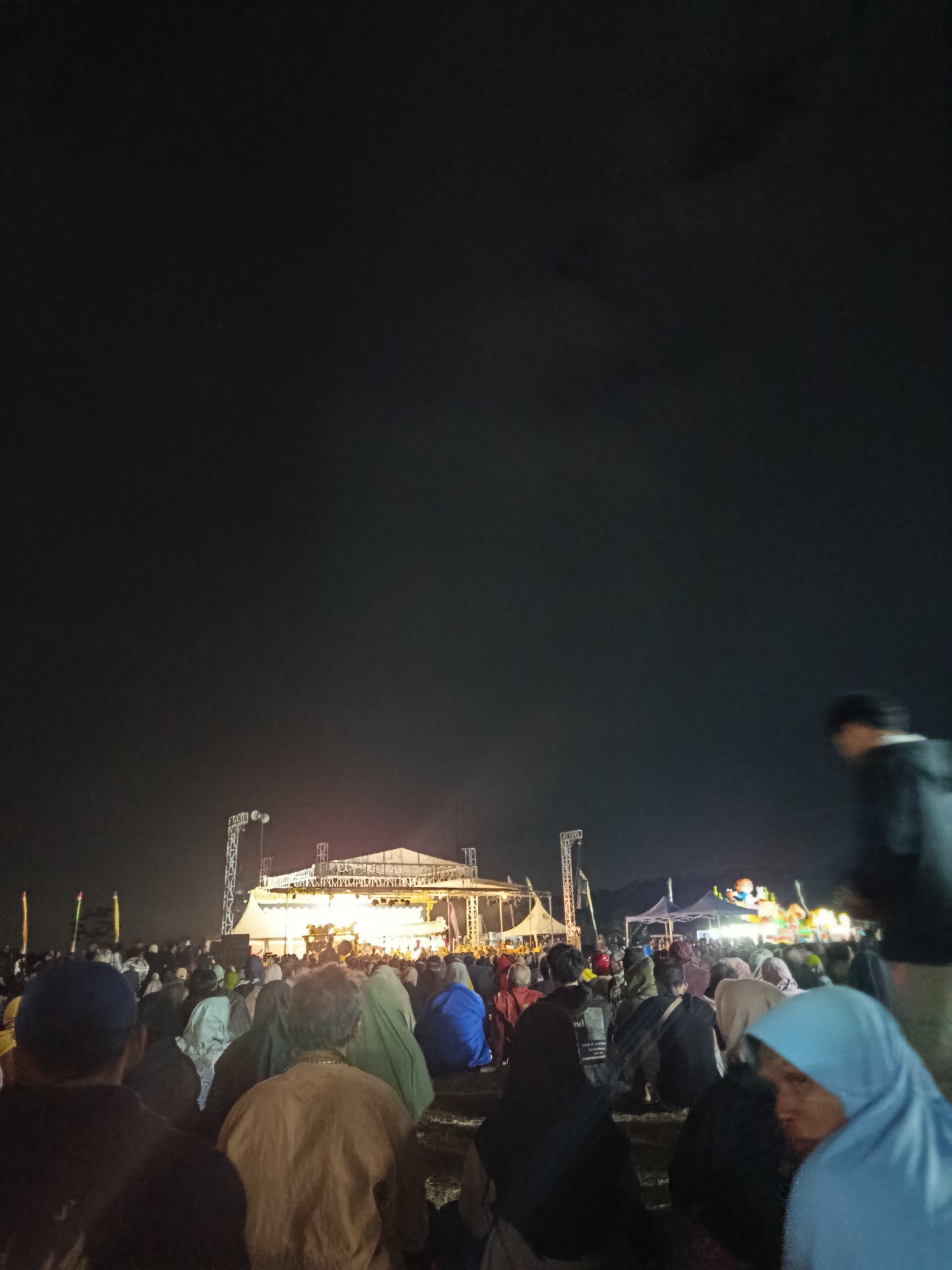The image depicts an outdoor evening event, likely a concert or festival, taking place in the city of Panembangan, Indonesia. A large crowd of people are gathered, seated on the ground, facing a brightly lit stage. The stage is elevated and has a large white tent-like structure covering it. Above and around the stage, there are scaffolding structures with lights, suggesting a professional setup. In the background, there are tents, possibly for vendors or other facilities, and the dark expanse of the night sky. A few individuals are visible in the foreground, with one person in the upper right corner walking away from the camera, partially blurred. The overall atmosphere appears to be one of communal gathering and entertainment.