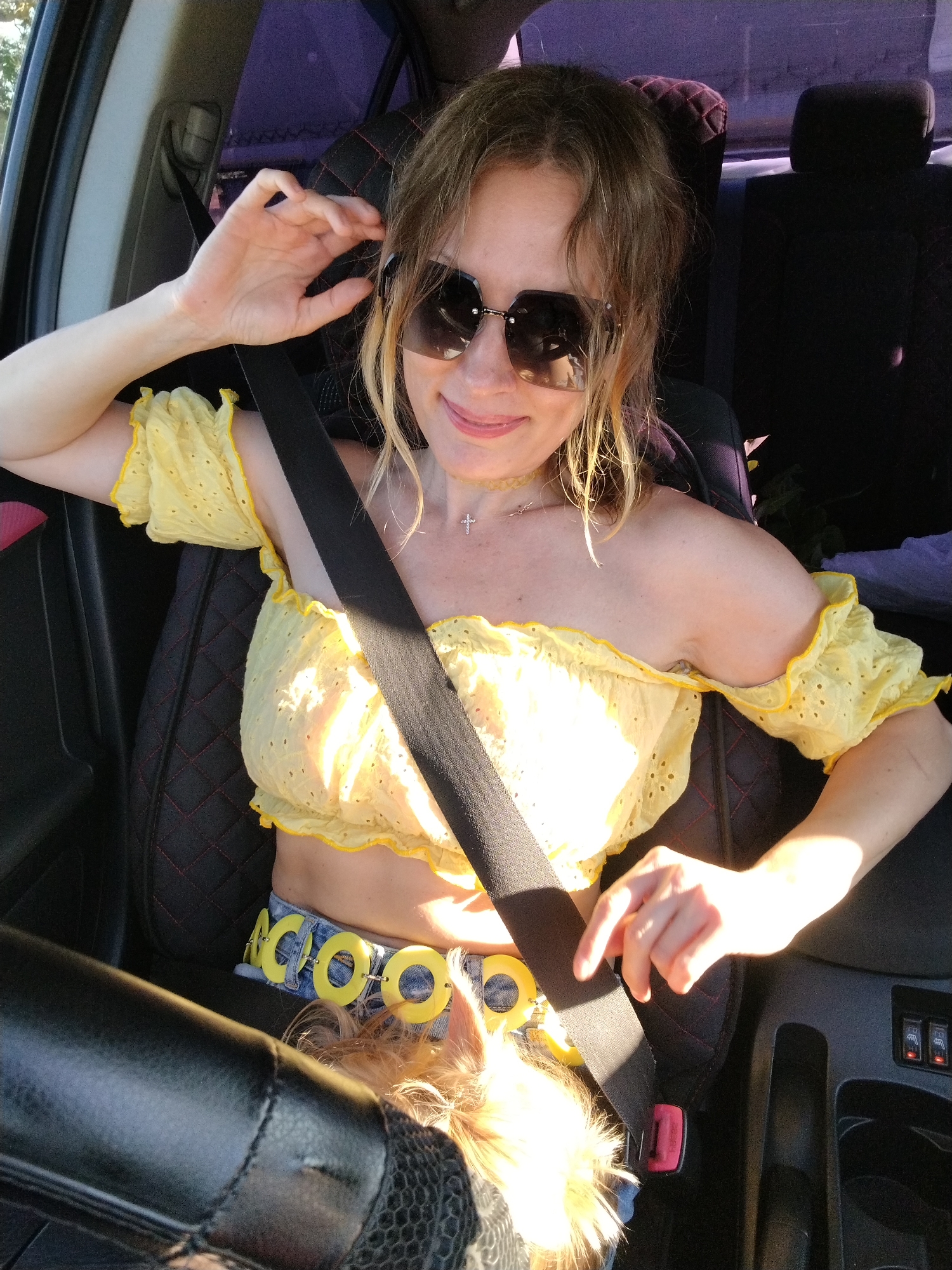 A woman with blonde hair and sunglasses is sitting in the driver's seat of a car, wearing a yellow off-the-shoulder top and a denim skirt with a yellow circular belt. She is adjusting her hair and has her seatbelt on. The interior of the car is visible, with a black leather steering wheel and car seats. The windows are tinted purple. The image appears to have been taken in Severskaya, Russia.