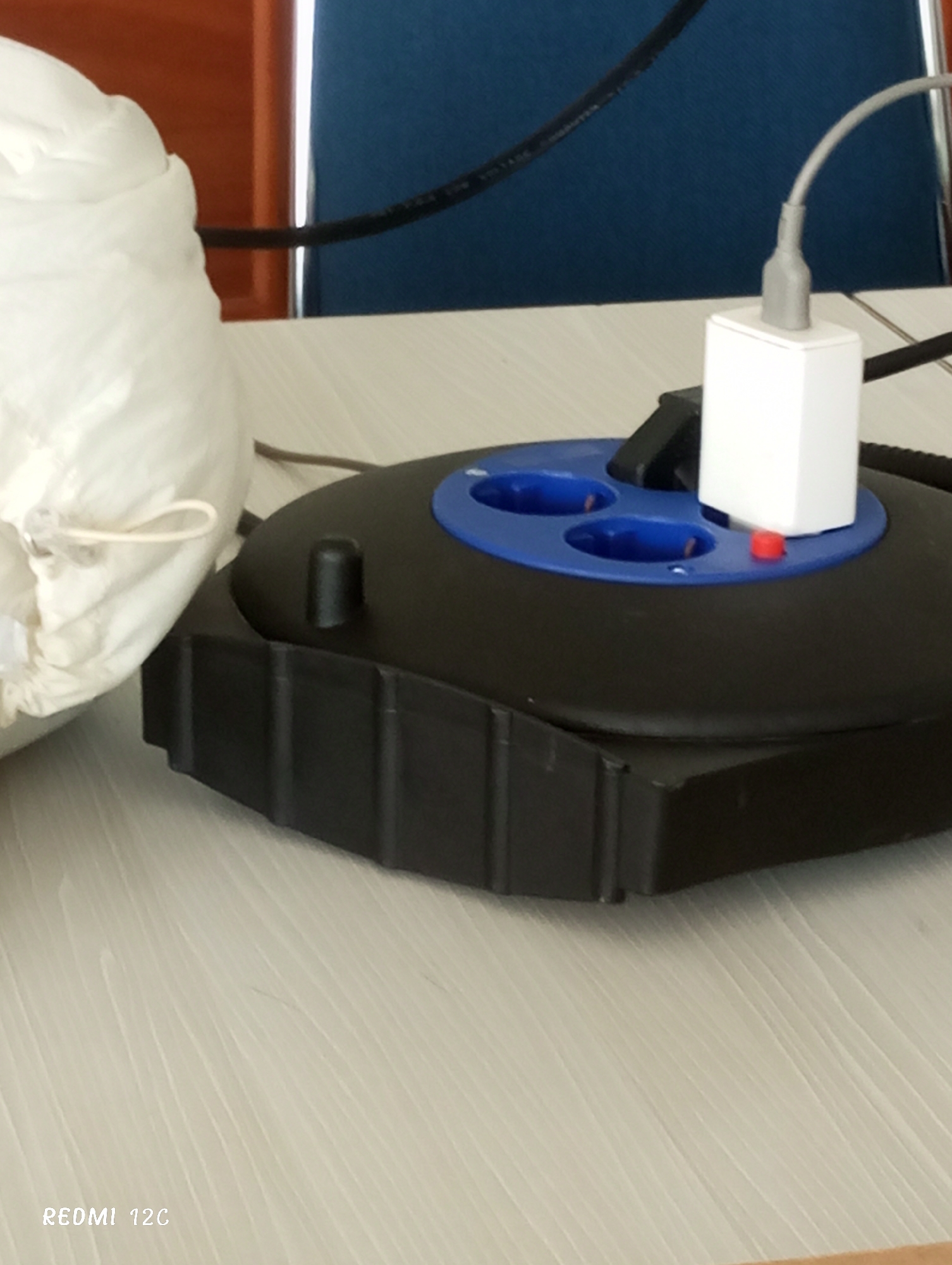 The image shows a black, round power strip with blue sockets sitting on a light-colored, possibly wooden, table. A white power adapter is plugged into one of the sockets. A white, cloth bag with a drawstring is partially visible to the left of the power strip. In the background, there is a blue chair and a wooden structure. The "REDMI 12C" watermark is visible in the bottom left corner. The image does not contain any information about the city of Lhokseumawe, Indonesia. There are no people visible in the picture.