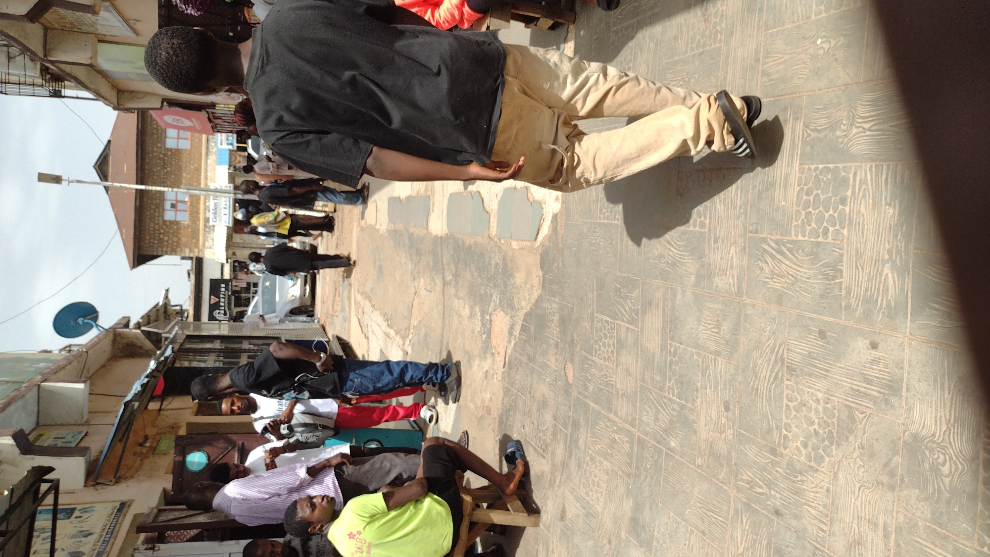 The image shows a street scene in Jos, Nigeria, with several people present. The street is paved with patterned tiles. Buildings line both sides of the street, and a utility pole with a satellite dish attached is visible on the left. Several people are standing or sitting along the left side of the street. One person is wearing a bright green shirt, and others are wearing casual clothing. A man in a black shirt and khaki pants is walking away from the camera along the right side of the street. Other individuals can be seen further down the street, and a white car is visible parked along the road. The sky is overcast.