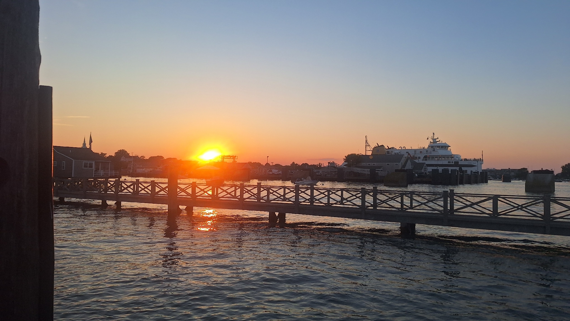 At sunset on Nantucket, a wooden pier extends across the water, its railings reflecting the warm glow of the setting sun. The sky is a gradient of orange and blue, with the sun's rays creating a shimmering path across the rippling water. In the distance, a large ferry boat is docked, its structure illuminated by the soft light. Along the shore, buildings with distinctive architecture can be seen, hinting at a coastal New England town. The overall scene evokes a sense of tranquility and the end of a day.