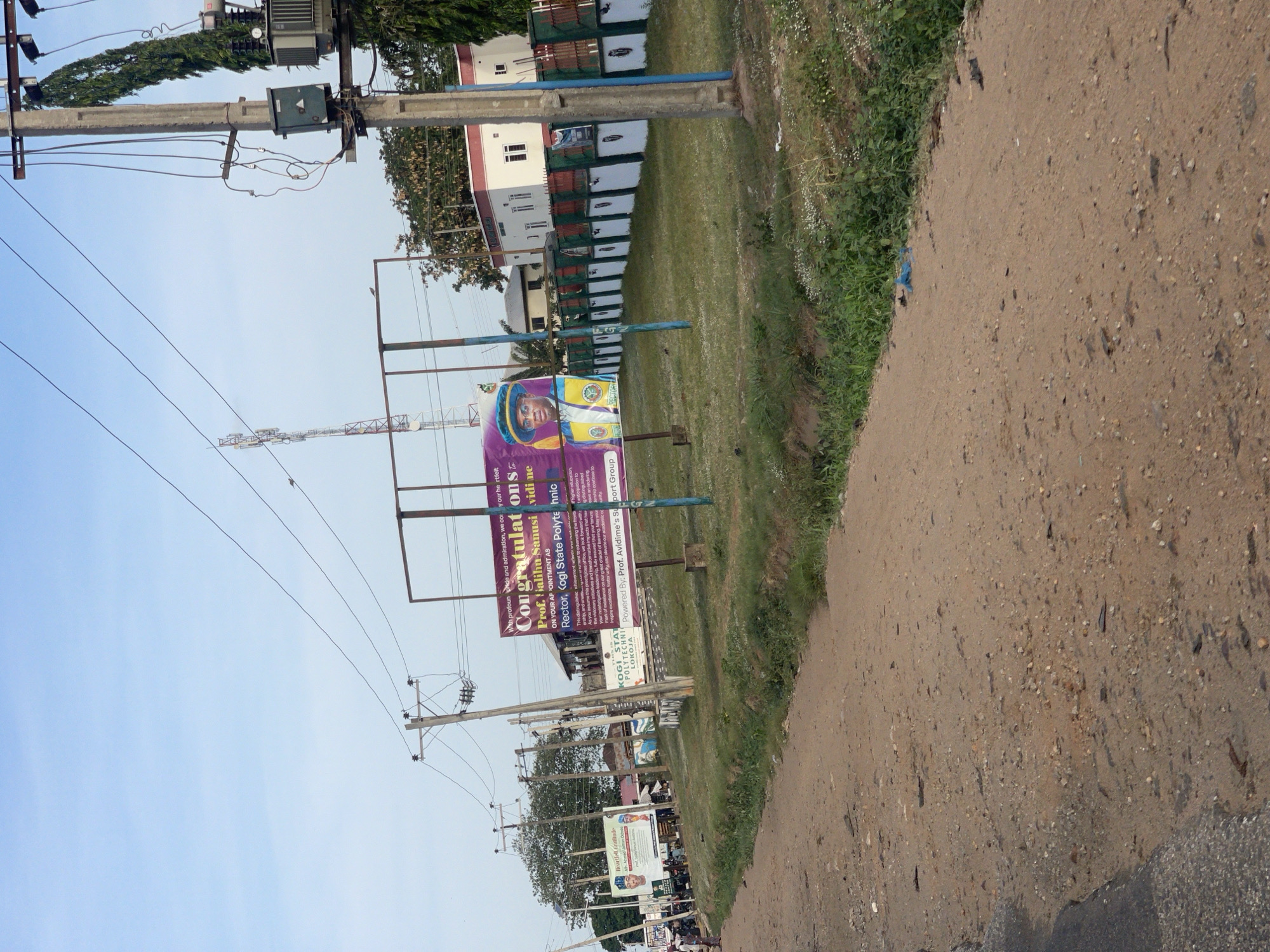 The image shows a dirt road with a grassy strip alongside it. A sign congratulating Prof. Galihu Sanusi on their appointment as Rector of Kogi State Polytechnic is visible. The location is in Lokoja, Nigeria. The sign is powered by Prof. Avidime's Support Group. In the background, there are buildings, trees, and power lines. Further in the distance, there are what appear to be more signs, with people gathering nearby.