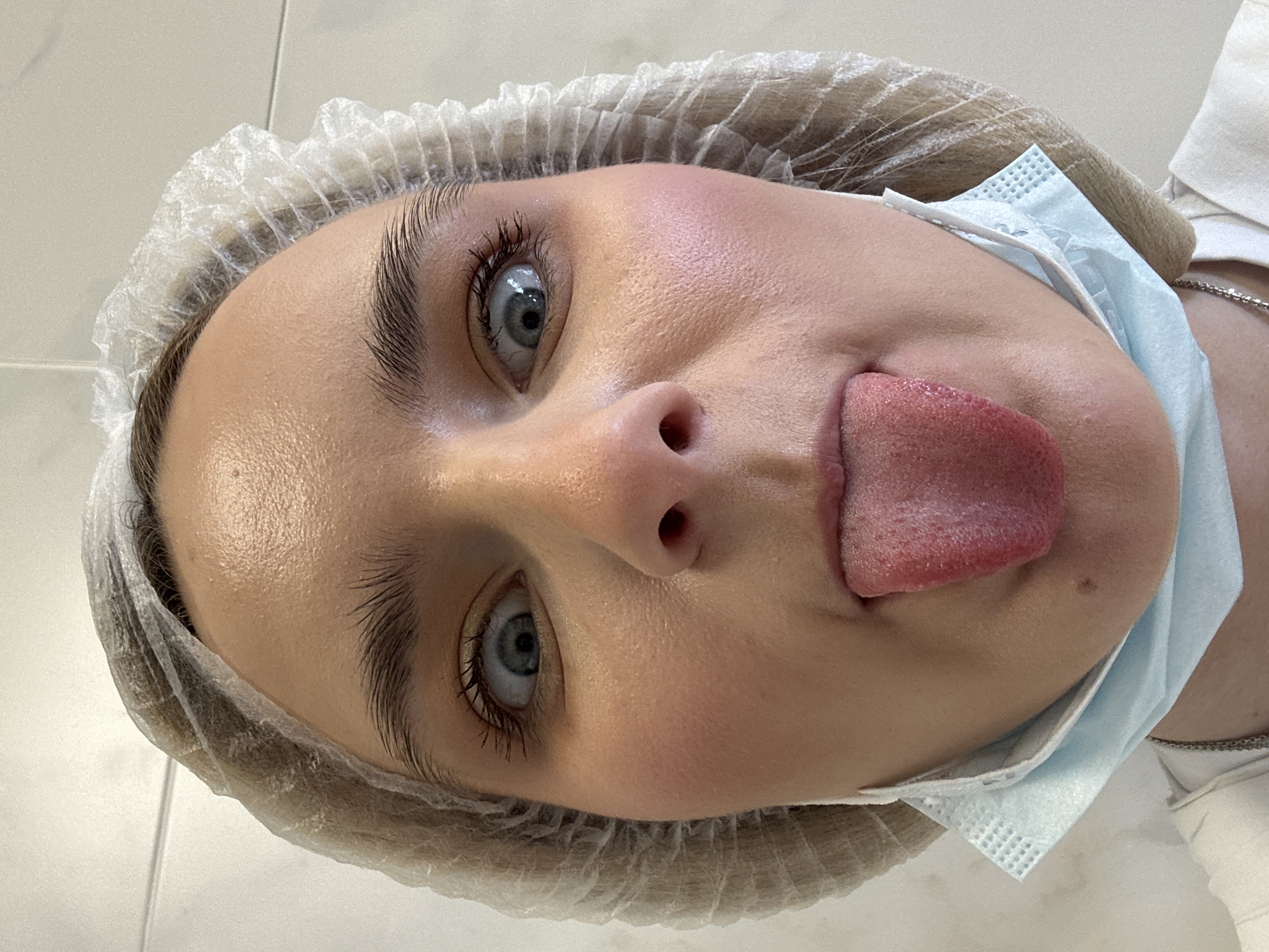 A light-skinned young woman is captured in a close-up, angled view. She is wearing a disposable surgical cap over her head and a light blue surgical mask pulled down below her chin, revealing her open mouth with her tongue sticking out. Her eyes are wide and light-colored, and her eyebrows are thick and prominent. The skin appears to be fair and smooth. A silver chain is visible around her neck. The background is a plain white surface. There is no information to suggest the picture was taken in St. Petersburg, Russia.