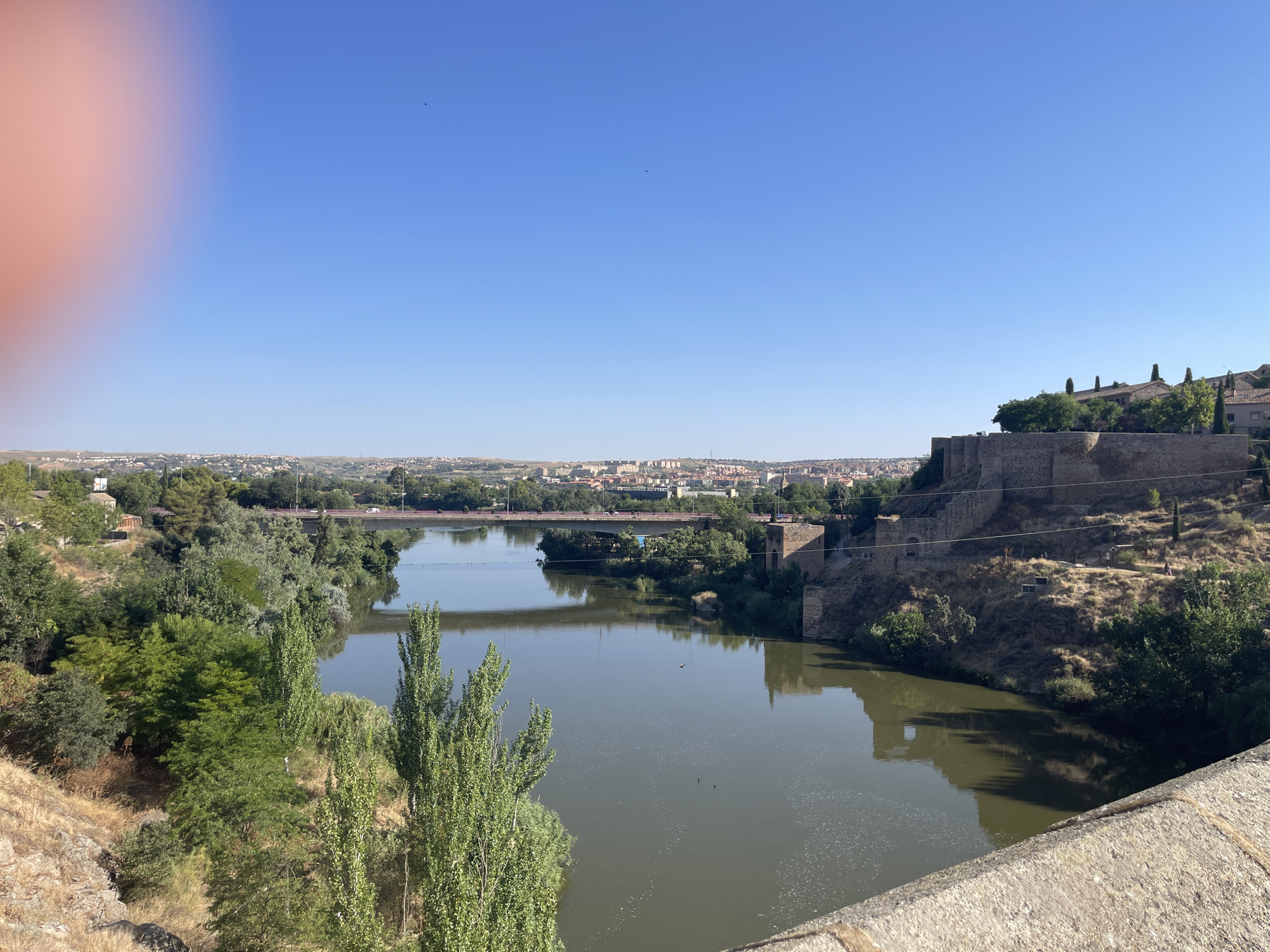 A wide shot captures the Tagus River flowing through the city of Toledo, Spain, under a clear blue sky. On the right bank, a stone fortification with battlements overlooks the river, leading to a hillside dotted with trees and buildings. Across the river, the city of Toledo sprawls across the horizon, with numerous buildings and a distant bridge carrying traffic. Lush green trees line the riverbanks on the left, with a rocky, sun-drenched slope in the foreground. The water reflects the sky and surrounding landscape. A faint reddish blur, likely a finger, is visible in the upper left corner of the frame.