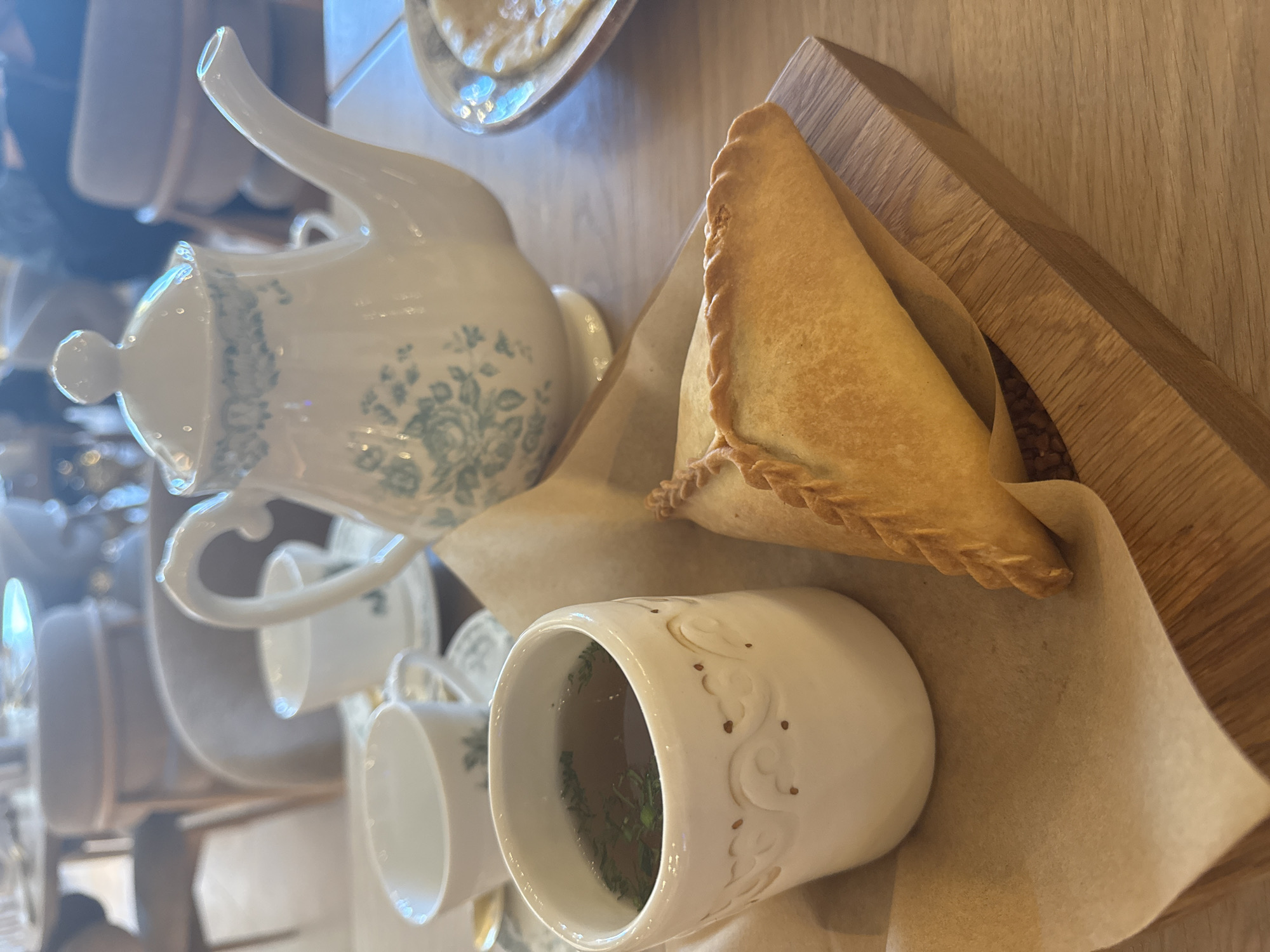 The image shows a table setting in Kazan, Russia, featuring a white teapot with a blue floral design, several white teacups, a cup of tea with green herbs, and a pastry on a wooden serving board with parchment paper. The table is likely in a restaurant or cafe.