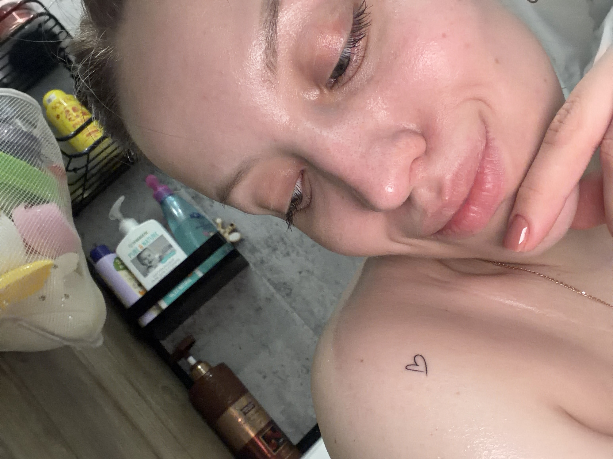 The image shows a close-up of a woman's face and upper chest. She has fair skin, and her face is tilted at an angle, with her eyes looking downward. Her right index finger is visible near her neck. A small heart tattoo is located on her chest. A thin gold chain is visible around her neck.
In the background, a shower caddy is seen holding various toiletries. These include a netted sponge bag, bottles of what appear to be shampoo and body wash, including one labeled "Pour & Nature" with a baby's face on the label. The background also shows gray flooring and a metal rack.
There is no information available within the image to suggest it was taken in the city of Reshetnikovo, Russia. The location cannot be definitively determined based on the visual content provided.