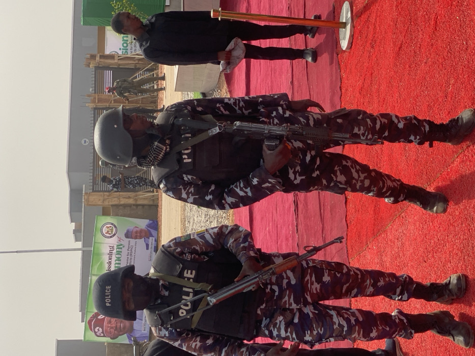 This image depicts an outdoor public event in Ifira, Nigeria. In the foreground, two armed Nigerian police officers stand on an extensive red carpet. The officer on the left wears a dark helmet with "POLICE," a black balaclava, a black tactical vest with "POLICE," a blue/white/brown camouflage uniform, and dark boots, carrying an AK-style rifle with a wooden stock. The officer on the right is similarly equipped with a dark helmet, a patterned scarf, a tactical vest, camouflage uniform, and dark boots, holding an AK-style rifle with a black stock.
In the mid-ground, other uniformed personnel, including additional police officers in camouflage and an individual in a red uniform, are visible near a metal gate. A civilian male in a black shirt and dark trousers stands to the right, holding a white patterned cloth. The background features two large green banners indicating a "COMMISSIONING OF Scheme for Person with Displacements (RSPIC)," displaying images of Nigerian politicians, specifically Senator Ahmed Lawan and His Excellency, Rt. Hon. (Dr) Aminu Bello Masari. The banner also clearly shows "Ifira." Behind the banners, several concrete buildings, possibly under construction or recently completed, are visible under a bright, clear sky. Gold-colored stanchions mark the event area on the red carpet.