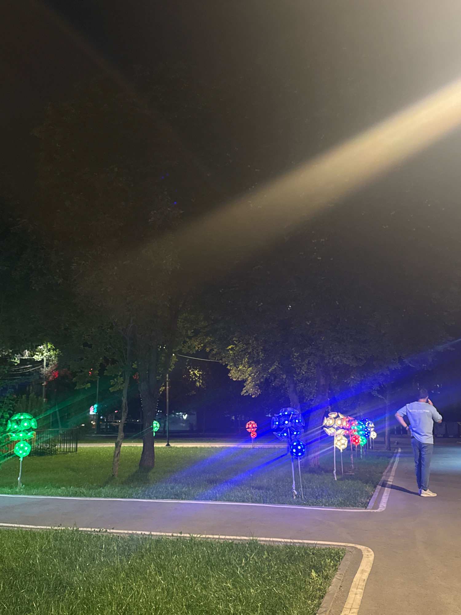 The image depicts a nighttime scene in Bishkek, Kyrgyzstan, possibly a park or public area, featuring decorative illuminated spherical objects placed along a paved pathway. A man in a blue shirt and jeans is standing on the pathway, looking away from the camera and holding a phone to his ear. The illuminated spheres are of various colors, including green, red, blue, and white, and are mounted on poles. Lush green grass borders the pathway on the left, and trees with dark foliage are visible in the background, partially illuminated by the lights. A bright beam of light cuts across the upper portion of the image from the top right. The overall atmosphere is serene and festive, with the colorful lights creating a striking contrast against the dark night sky.