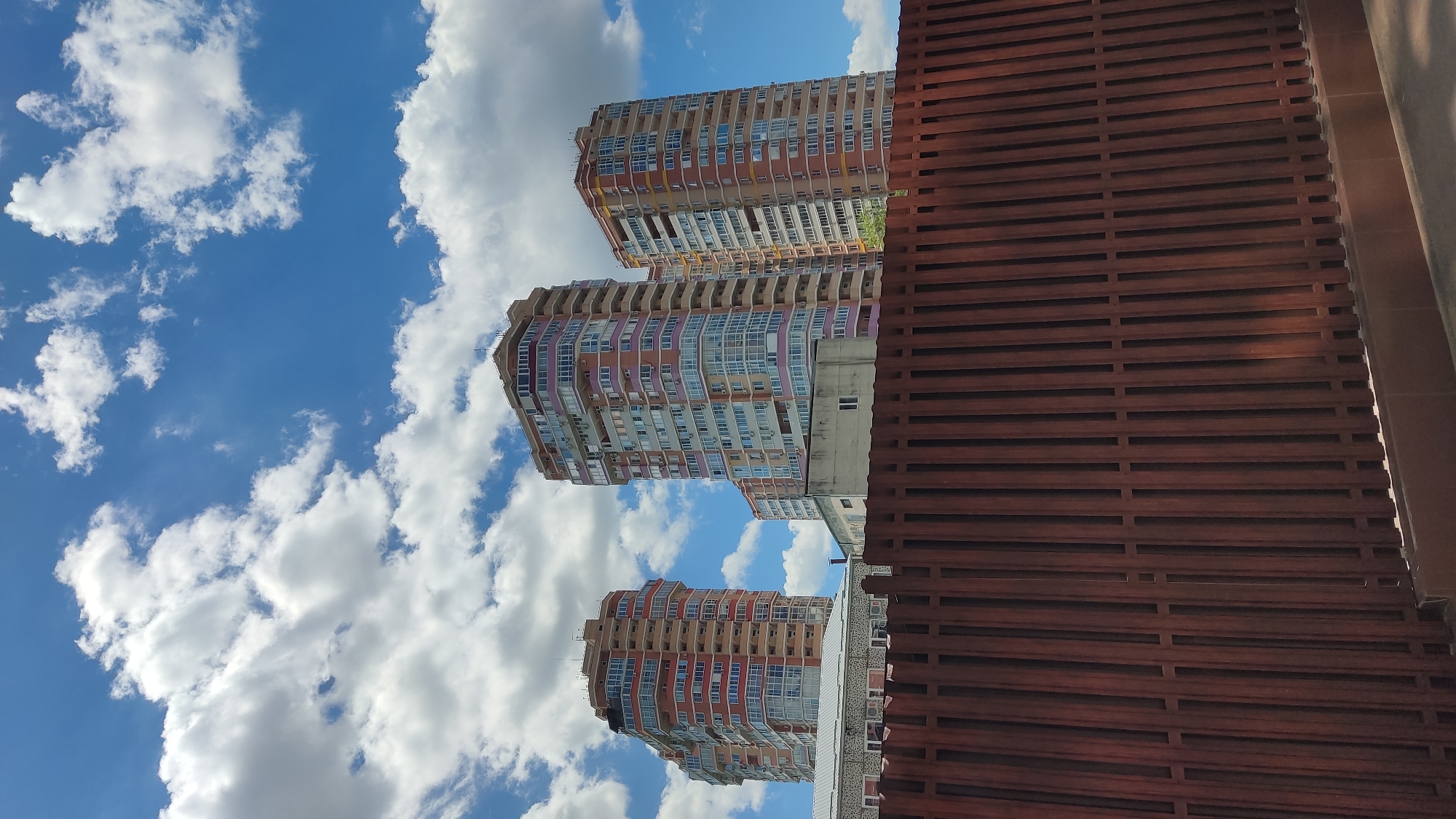 The image captures a view of modern high-rise apartment buildings under a bright blue sky dotted with fluffy white clouds. The buildings, located in the city of Krasnodar, Russia, feature colorful facades with balconies and numerous windows. In the foreground, a dark brown, vertical slat fence runs across the width of the image, partially obscuring the lower portion of the buildings and a smaller building situated between the fence and the high-rises. The overall scene suggests a daytime urban environment.