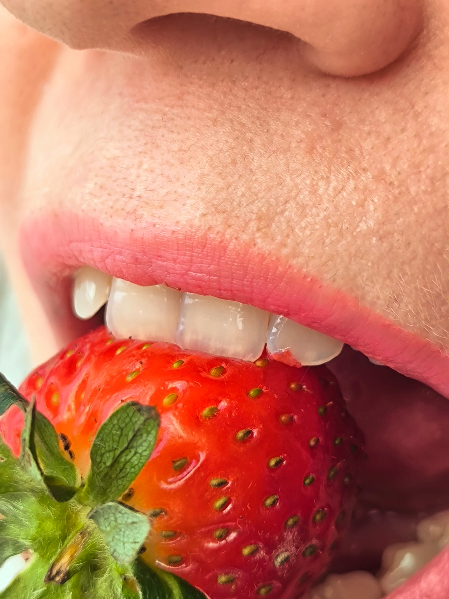 A close-up shot features a person's mouth biting into a strawberry. The lips are pink, with visible texture and skin details. The person's teeth are slightly visible, pressing into the strawberry's surface. The strawberry is red and has green leaves at the top, indicating it is fresh. The seeds on the strawberry's surface are noticeable. The background is out of focus, appearing to be skin. The location in the city of Unknown, Unknown, is indeterminate.