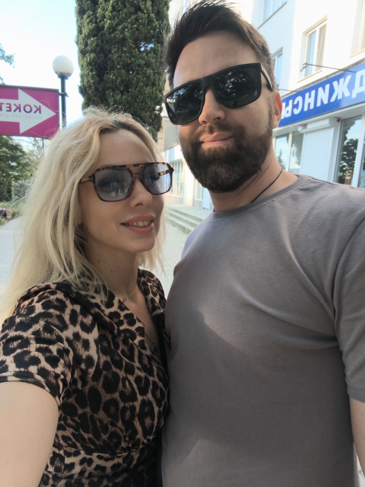In Sevastopol, Ukraine, a couple poses for a selfie outdoors. The man, with a beard and sunglasses, wears a grey t-shirt. The woman, with blonde hair and sunglasses, is dressed in a leopard-print top. Behind them, a building with blue signage and an arrow-shaped sign with Cyrillic text indicating "KOKEL" are visible. A tall evergreen tree stands to the left. The scene appears to be a street in a city.