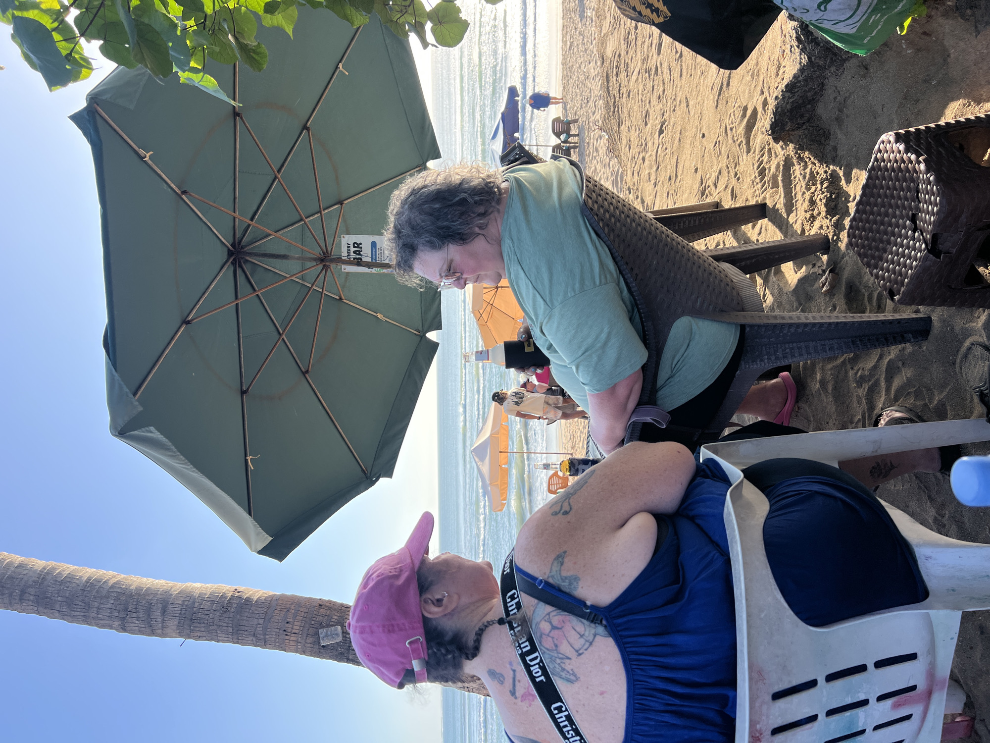 Two women are seated at tables on a sandy beach in Kuta, Indonesia, near the ocean. One woman, wearing a pink baseball cap and a blue dress, is seated in a white plastic chair and has tattoos on her back and arms. The other woman, wearing a green t-shirt and glasses, is seated in a dark brown wicker chair. Both women are facing away from the camera and appear to be engaged in conversation. An umbrella provides shade over the seating area, and a sign that reads "CHERRY BAR" is visible on the underside of the umbrella. The ocean is visible in the background, with a few people walking along the shore and a small orange umbrella in the distance. Palm trees are also present in the scene, indicating a tropical environment.