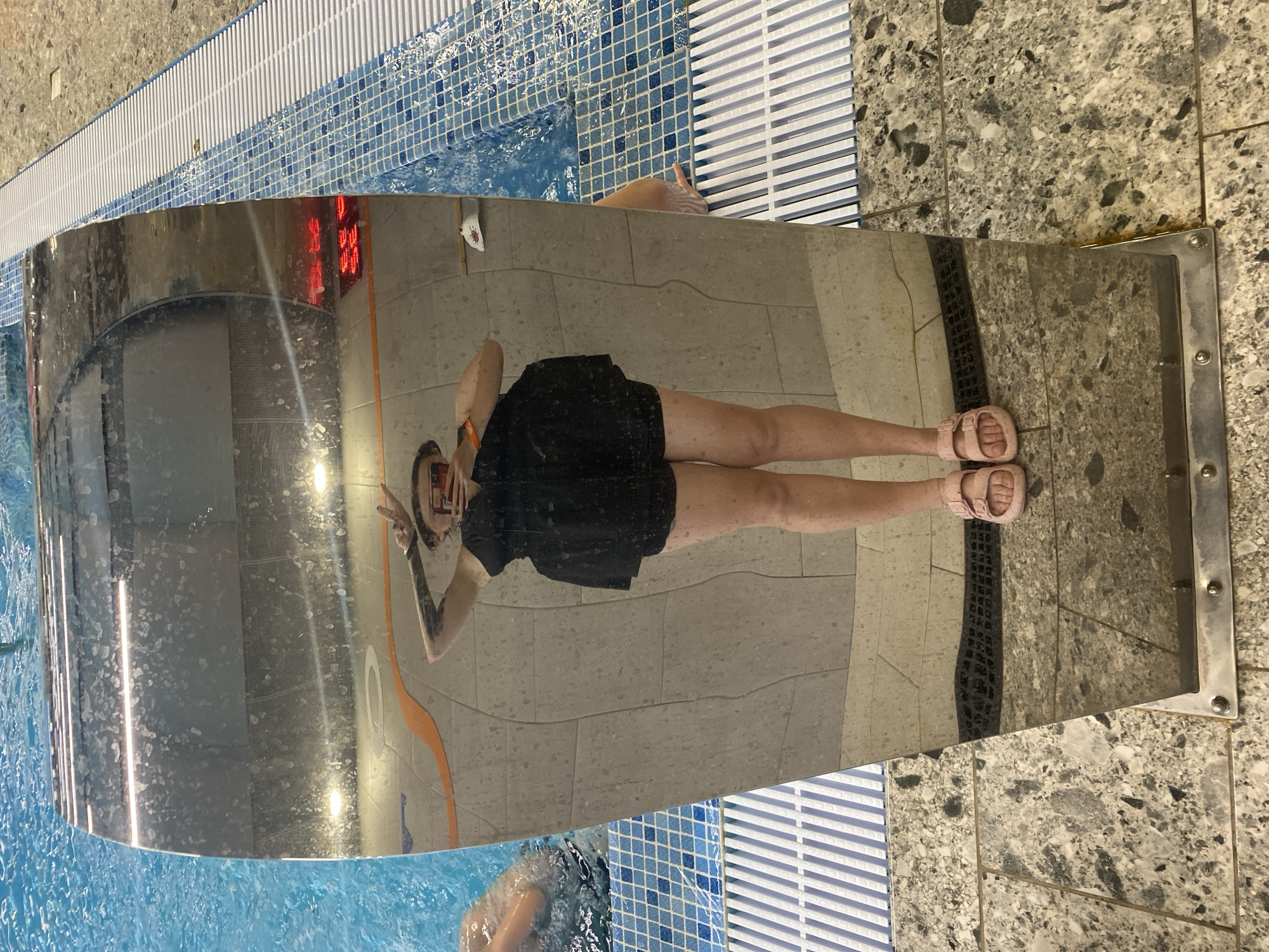 A full-body mirror reflection of a person wearing a black dress and pink sandals, standing by a swimming pool. The person is holding up a phone to take the picture and has tattoos on their arms. The background shows a tiled wall, the blue water of the pool, and a grate. The mirror is curved, distorting the reflection. The location is the swimming pool area of the Votkinsk city maternity hospital in Votkinsk, Russia, as indicated by the digital clock displaying "01:05" and the red temperature display that can be seen on the wall in the reflection of the mirror.