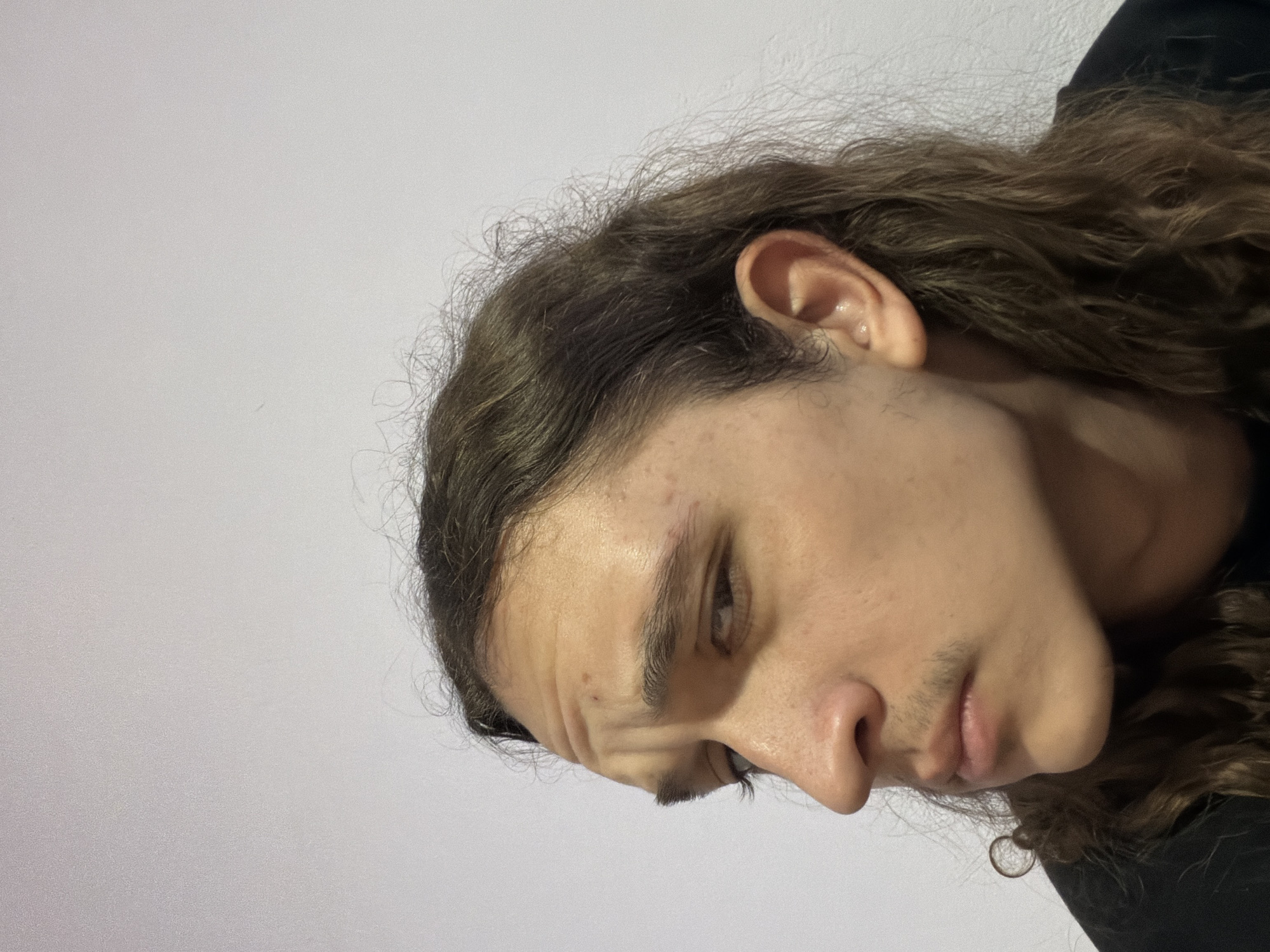 A close-up, medium shot depicts the right side of a person's face, angled downwards. The individual has medium-length, curly brown hair and fair skin. Details include an ear, an eye, a nose, and a mouth. The person is wearing a dark-colored garment. The background is a plain, light grey surface. There is no information suggesting the photo was taken in Asenovgrad, Bulgaria, or any identifiable location.