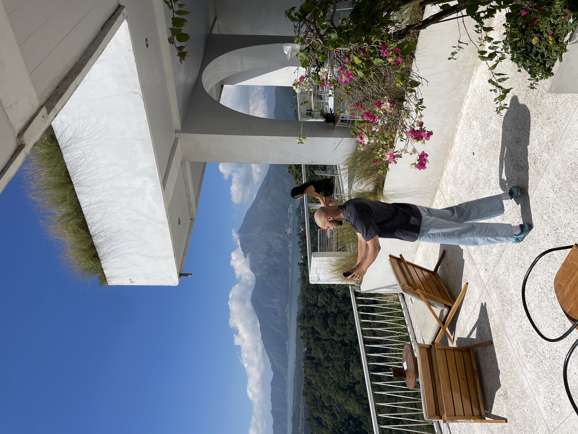 A bald man in a dark t-shirt and light blue pants is standing on a terrace in Kintamani, Indonesia, taking a selfie with his phone. He is facing away from the camera, looking at his phone. To his left, a woman in a black dress is sitting in a chair looking out at the view. The terrace overlooks a lush green valley with a large lake and mountains in the distance. The architecture is modern with white concrete walls and a white railing. There are wooden chairs and tables on the terrace. Pink and white bougainvillea flowers are blooming in the foreground. The sky is clear blue with a few scattered clouds.