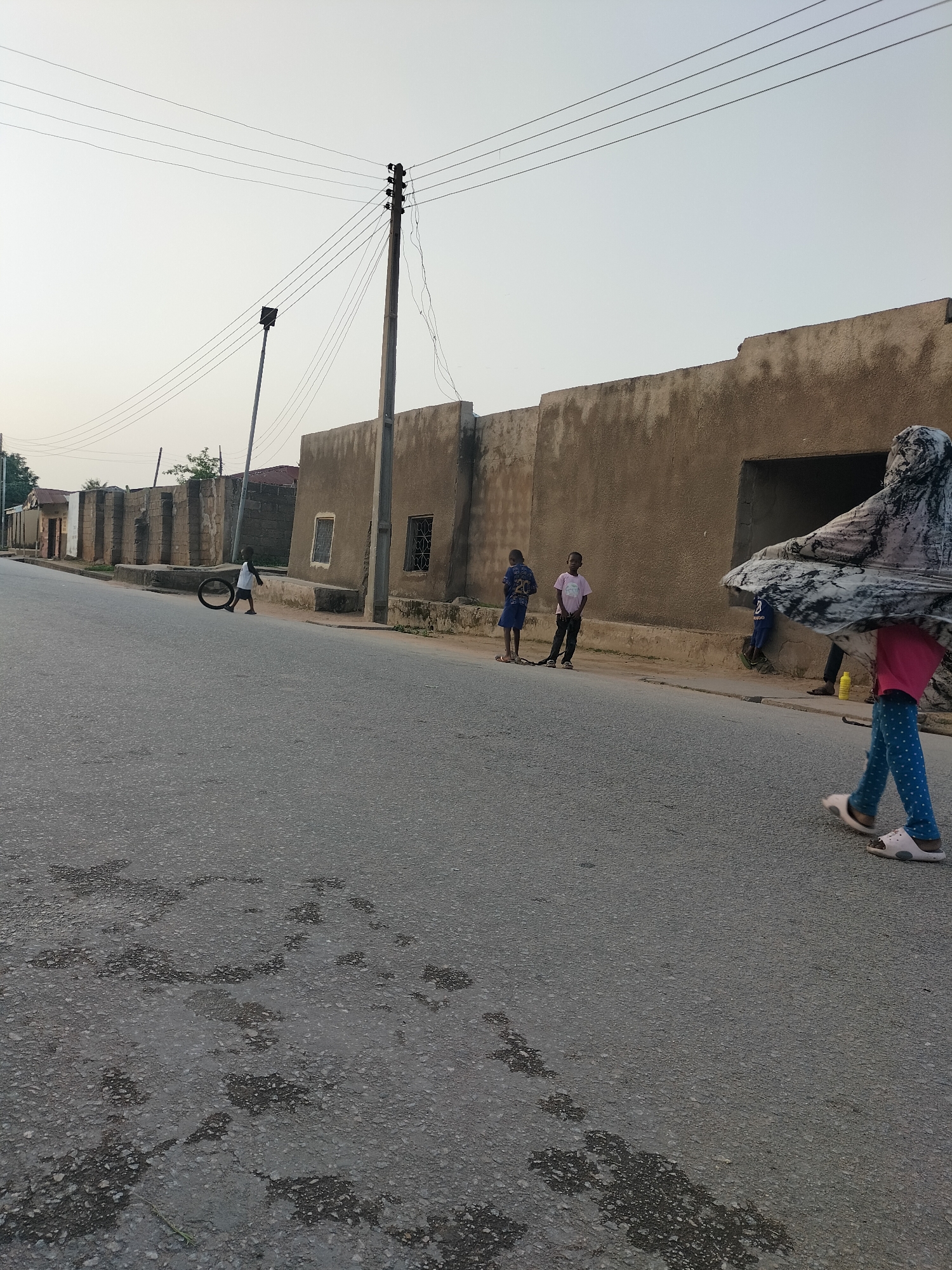 The image depicts a street scene in Jalingo, Nigeria, likely during daytime hours given the illumination. The foreground is dominated by an asphalt road, showing some signs of wear and tear with darker, damp-looking patches. A young child is visible on the left side of the road, pushing a tire. Further down the street, on the right side of the road, a person is walking towards the camera, their lower body covered by a patterned fabric that appears to be part of a hijab or veil, and they are wearing blue polka-dot leggings and light-colored sandals. In the background, several children are standing near a low wall in front of a building. The buildings are constructed with rough, plastered walls, and some have visible windows with decorative bars. Overhead, electrical wires are suspended from a utility pole, and another pole with a street lamp is also present. The sky is overcast, suggesting a hazy or cloudy day. The overall impression is of a residential or semi-commercial area in a Nigerian city.