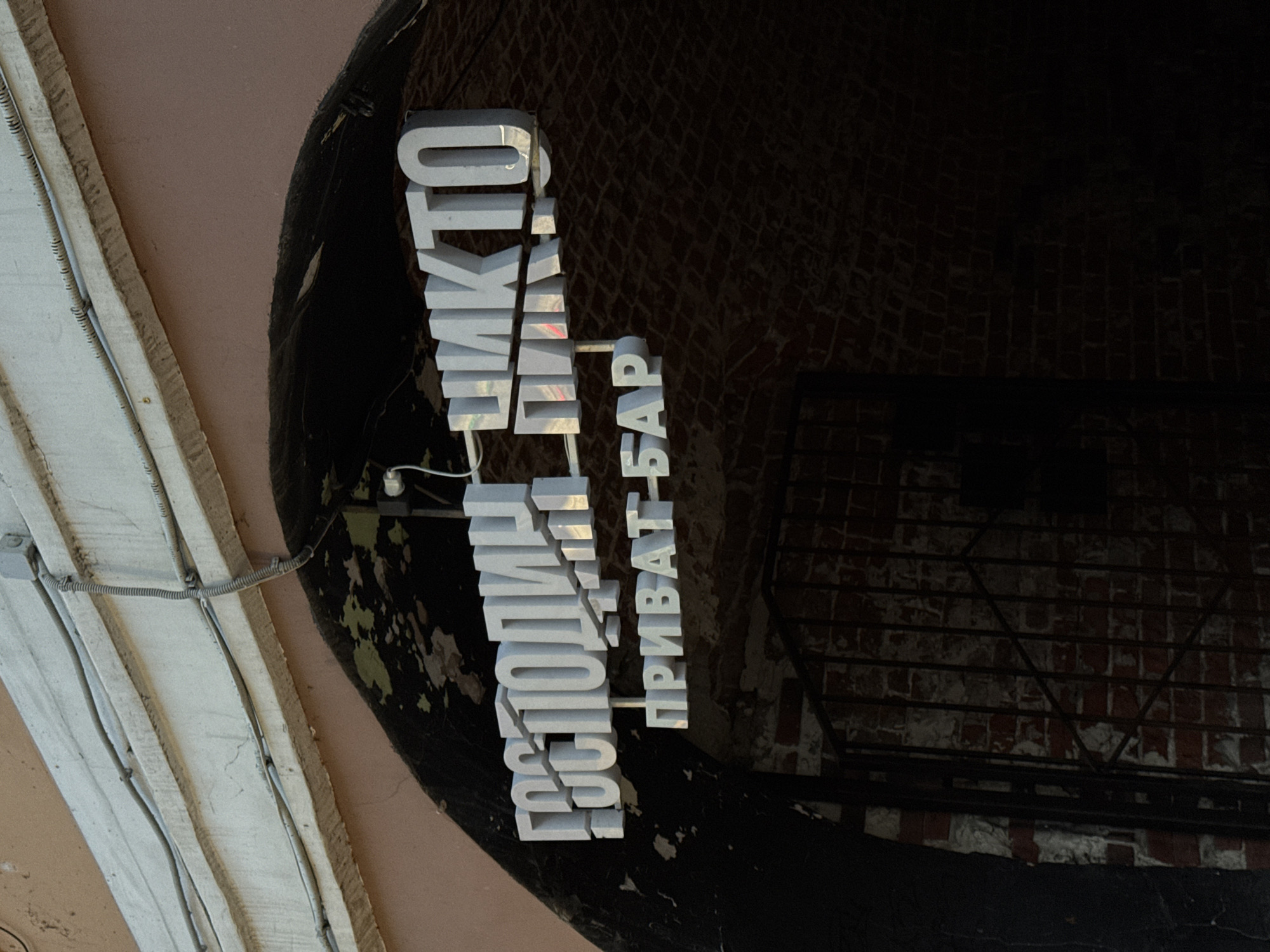 The image shows a sign in Russian, "ГОСПОДИН ЧИКИТО ПРИВАТ-БАР" ("Mr. Chiquito Private Bar"), affixed to a wall or entranceway. The sign is white and illuminated, with a three-dimensional effect. The background includes architectural elements, such as a vaulted ceiling and a brick wall, suggesting an interior or entryway. There are also electrical wires and conduits visible on the left side of the image. The location of the photo is the city of Nizhny Novgorod, Russia.