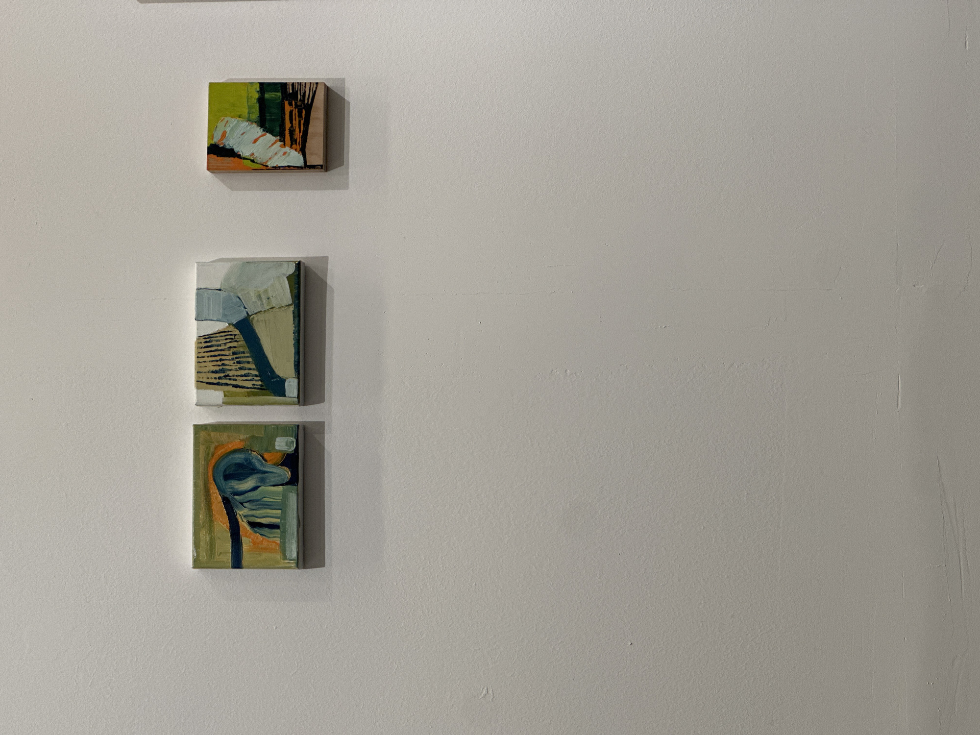 Three small abstract paintings hang vertically on a white wall. The top painting features green, white, and brown hues. The middle painting uses shades of grey, blue, and beige. The bottom painting has green, orange, and blue tones. The wall appears to be textured and slightly off-white. The location is not indicated.