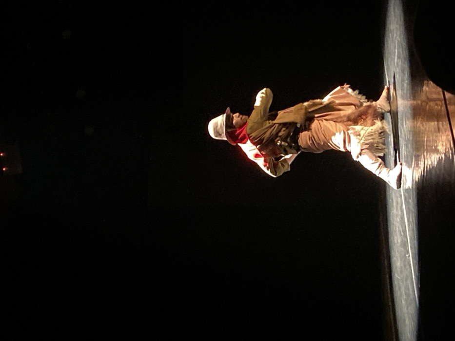 A single individual is depicted on a dark stage, illuminated by a focused spotlight. The person, appearing male, is captured in profile, facing right, in a dynamic pose suggestive of a dance or performance. The attire consists of a light-colored fedora-style hat, a red hooded garment covering the head and upper back, a light-colored long-sleeved shirt, and an olive-green poncho or shawl draped over the left shoulder and torso. The lower body is dressed in loose-fitting, light-colored trousers or a skirt-like garment featuring a fringed hem, paired with light-colored, soft footwear. The stage floor is dark and reflective, displaying a light-colored linear marking. The background is uniformly black, with two small red lights faintly visible in the upper central area. The specific venue within Buenos Aires, Argentina, is not identifiable from the visual evidence.