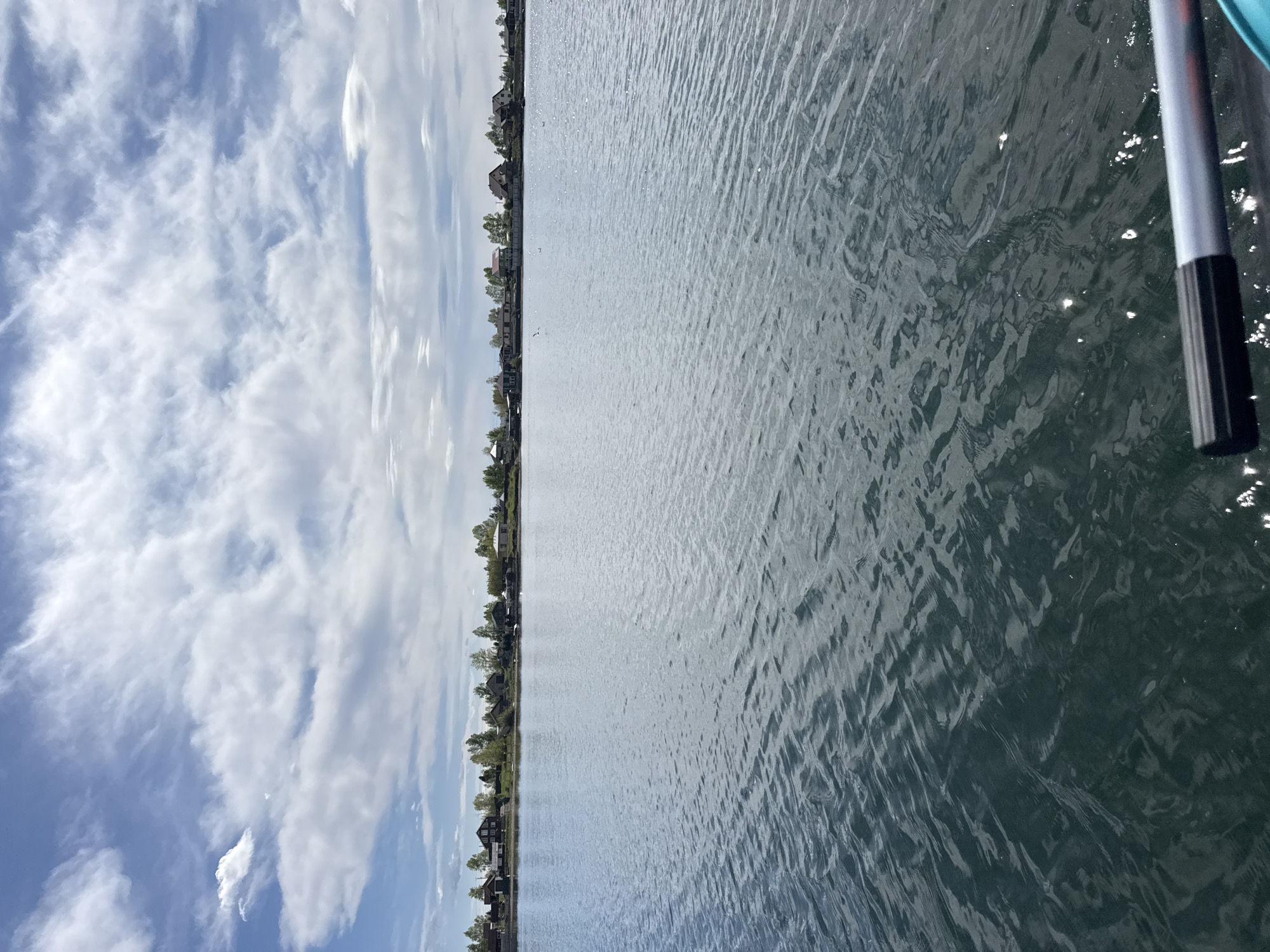 The image depicts a body of water, likely a lake or river, with a shoreline visible on the left. The sky is filled with scattered clouds. On the right side of the image, a portion of a paddle or oar is visible, suggesting the photo was taken from a boat or kayak. The water's surface shows slight ripples and reflections. Based on the presence of residential structures along the shoreline, and assuming the request for the city of Irkutsk, Russia is accurate, the image might depict a view of the Angara River or a lake in the vicinity of Irkutsk. However, without more information or identifiers, providing a specific location is challenging. The scene conveys a sense of tranquility and outdoor activity.