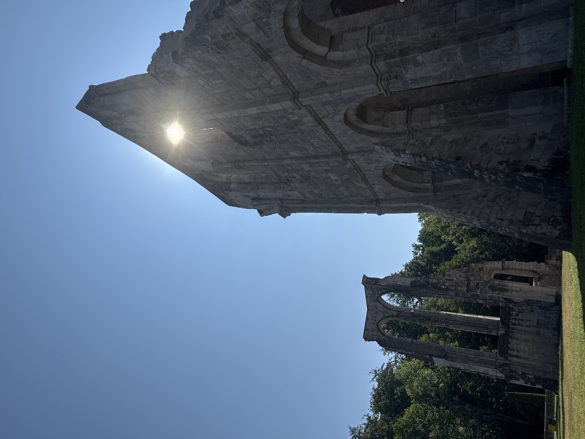 The image shows the ruins of the Walkenried Abbey in Walkenried, Germany. The abbey ruins are made of stone and are partially covered in moss. The ruins are set against a bright blue sky. The sun is shining brightly, and its rays are visible through a gap in one of the ruins. In the background, there are trees and a grassy field. The image is taken from a low angle, looking up at the ruins.