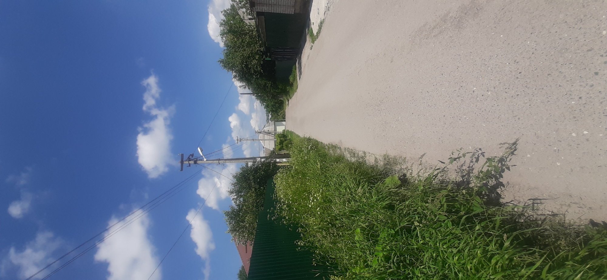 The image depicts a rural scene in Yessentukskaya, Russia, on a sunny day. The perspective is from a low angle, looking up and to the right. The sky is a vibrant blue with scattered white clouds. A power line pole with visible wires and a street light stands on the left side of the frame. To the right of the pole, a paved road stretches into the distance. The road is bordered by lush green grass and foliage. In the background, several houses with different rooflines and building materials are visible, along with trees. The overall impression is one of a tranquil, everyday summer day in a village. The lighting suggests it is daytime, likely mid-morning or afternoon, given the bright sunlight. No people are visible in the image. There is no visible text.