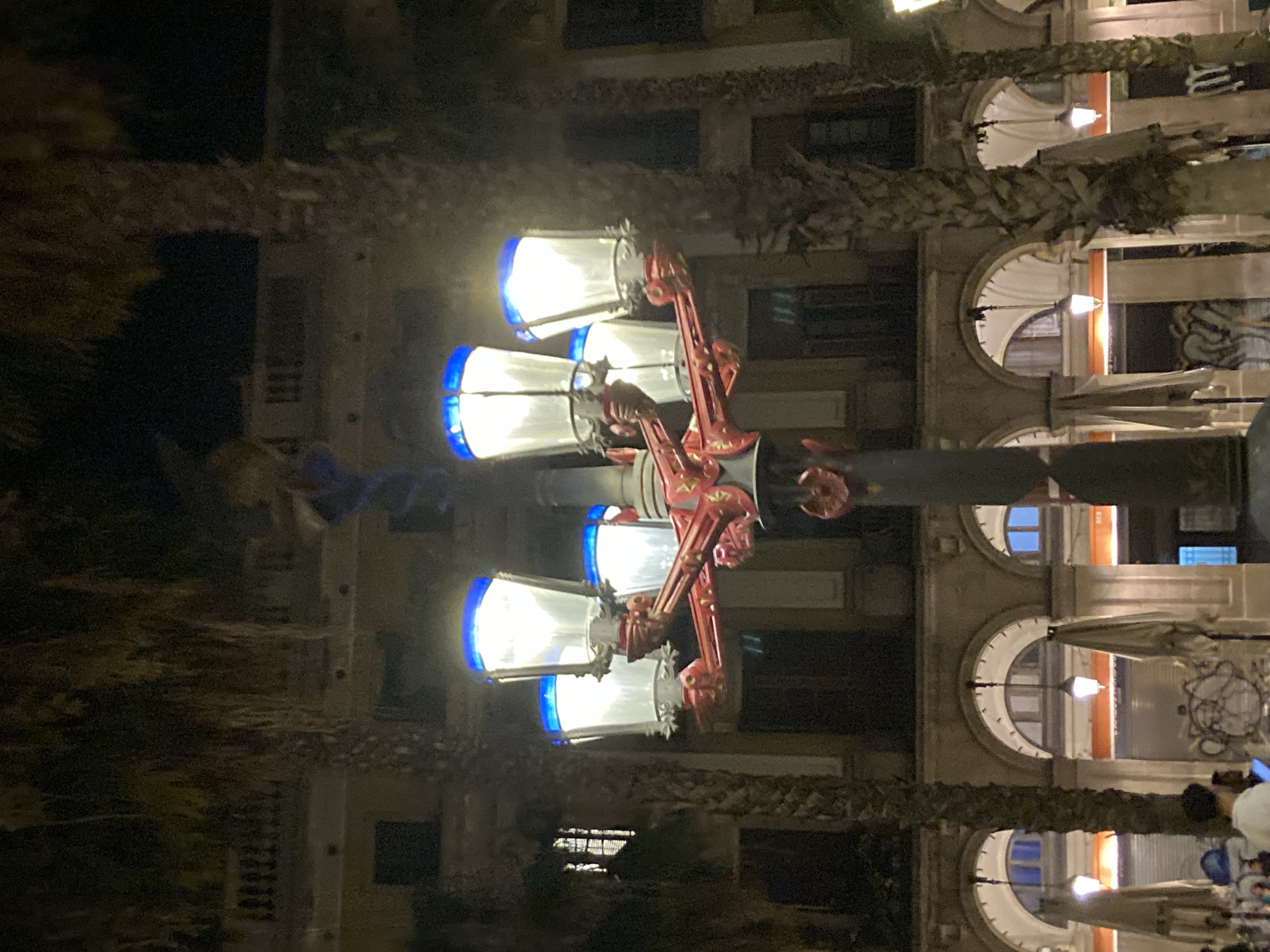 This image shows a lamppost with multiple lights and blue tops, in front of an arched building with palm trees. The lamppost is decorated with red metalwork. The building appears to be a commercial or residential building, with awnings and windows visible. The scene is lit by the lamppost, suggesting it is nighttime. The location is identified as Barcelona, Spain. People are visible in the foreground, blurred and out of focus, suggesting a public area. The overall impression is that of a street scene at night in a city.