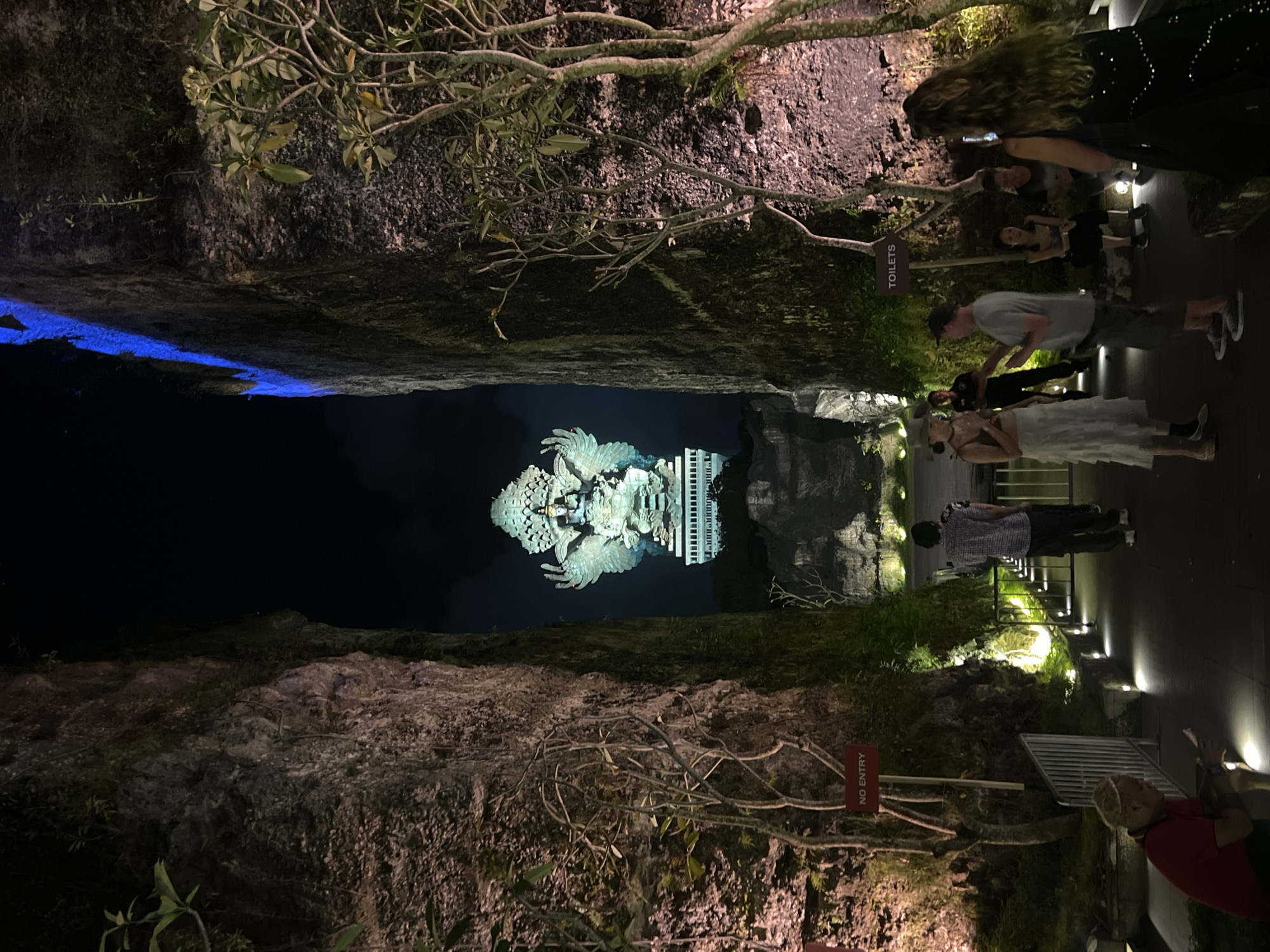 This nocturnal image captures a dramatic scene within a deeply cut gorge or quarry, illuminated by artificial lights. The perspective is from a viewing platform overlooking a body of dark water, which reflects a magnificent, brightly lit statue in the distance.
On the left side, a rugged rock face, sparsely covered with vegetation, descends towards the water. A striking blue light source illuminates a section of the water along this rock wall. The water itself appears dark and still, perfectly mirroring the central sculpture.
In the middle ground, across the water, stands a colossal, intricately detailed statue. It depicts a winged, bird-like figure, strongly resembling the mythical Garuda, carrying a human-like deity. The statue is predominantly light in color, glowing under focused illumination, and its reflection creates a symmetrical image on the water's surface. Behind the statue, some modern, illuminated building structures are visible.
On the right side of the image, a paved pathway runs alongside another textured rock wall, also adorned with sparse greenery. Several people, likely visitors or tourists, are present on this pathway. They are observing the statue or moving along the path. Among them, a woman with long hair in a dark dress appears to be looking at a phone, while another woman wears a distinctive white, tiered dress. A man in a grey t-shirt stands further back. The pathway is well-lit by ground-level fixtures.
Visible text includes a sign pole reading "TOILETS" and a red "NO ENTRY" sign affixed to the rock face on the right. The overall atmosphere suggests a tourist attraction or an event taking place at night, highlighting the grandeur of the statue and its unique setting.