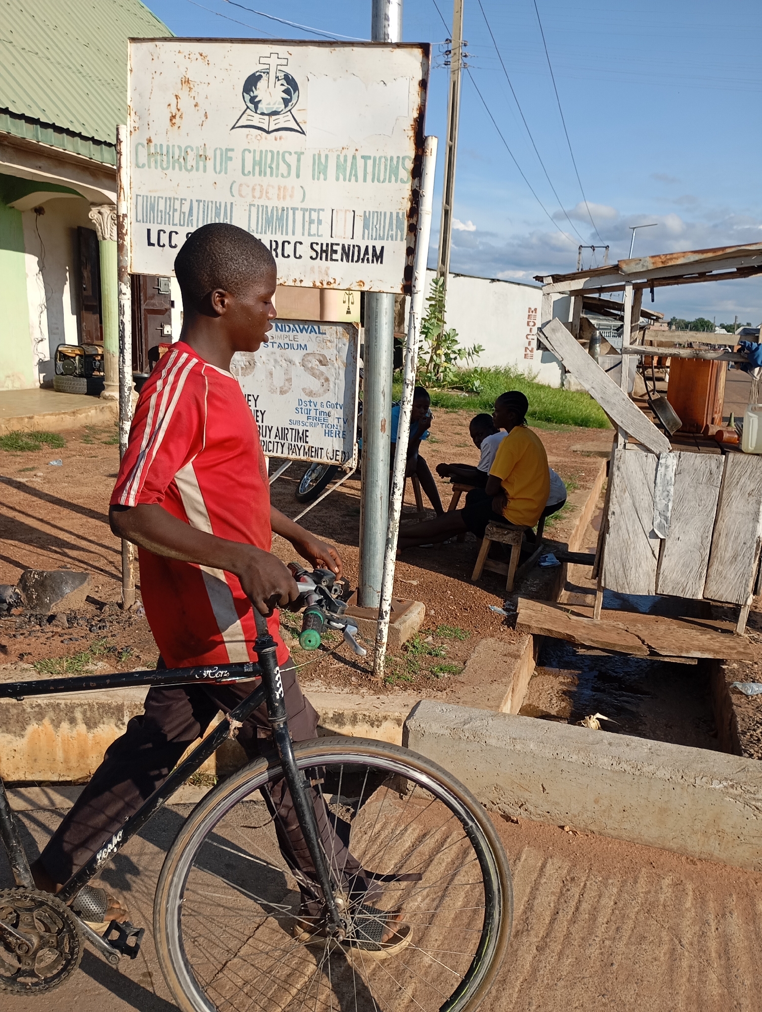 In the city of Shendam, Nigeria, a young person, dressed in a red and white striped shirt and dark pants, walks their black bicycle past a sign for the "CHURCH OF CHRIST IN NATIONS (COCIN) CONGREGATIONAL COMMITTEE LCC RCC Shendam." The sign, weathered and faded, is mounted on a pole near a building with a green roof. In the background, a few other individuals are seated on wooden stools, possibly at a roadside stall. The ground is a mixture of dirt and pavement, with a concrete structure and a small stream or drainage channel visible. The sky is blue with scattered clouds, indicating daytime.