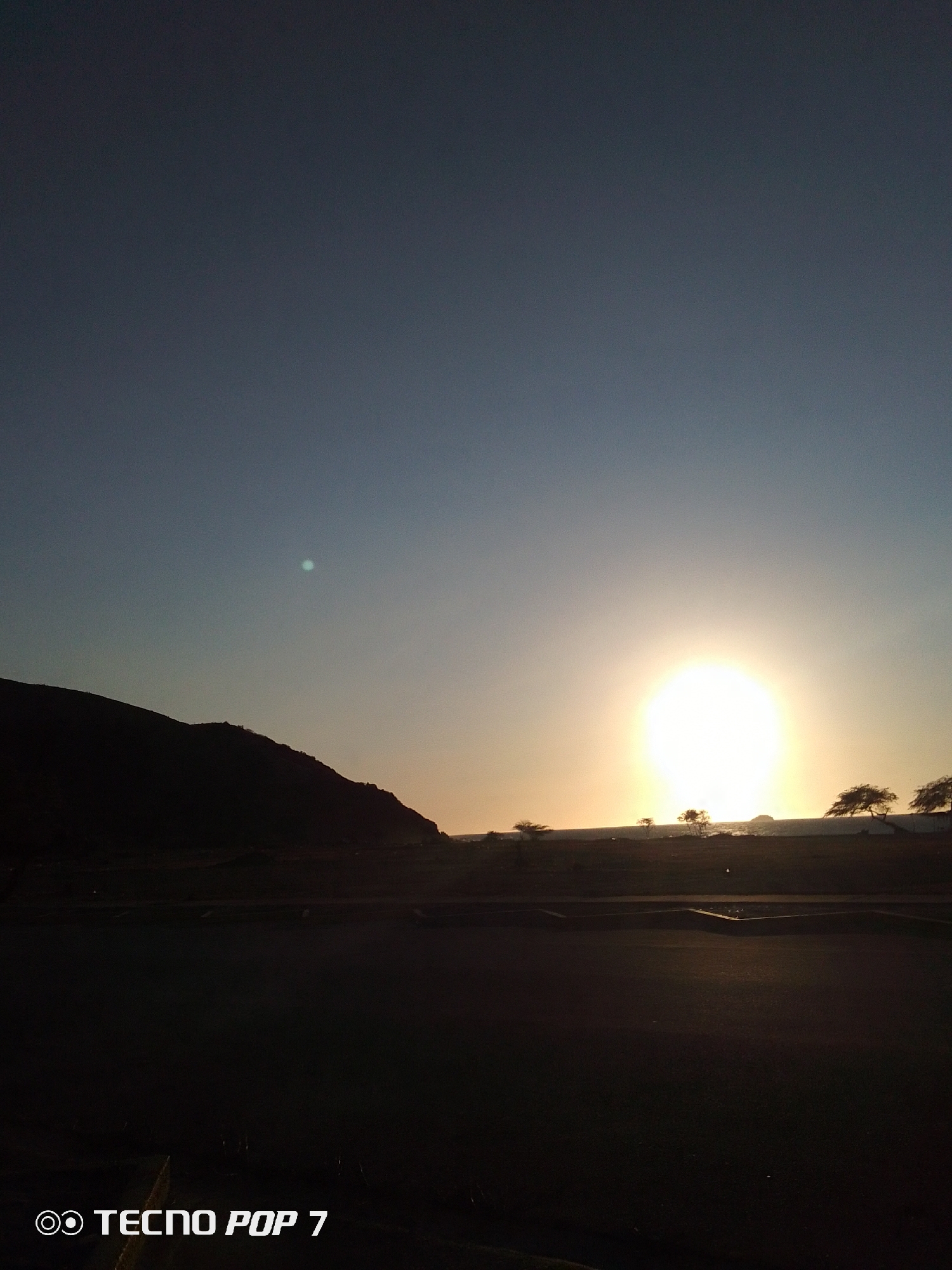 The image depicts a landscape at sunset. The sky transitions from a dark blue at the top to a lighter orange near the horizon. A bright sun is visible, partially obscured by the landscape. Silhouetted trees and a mountain range are present. The foreground is dark, obscuring details. The location is Unknown, Unknown. The presence of the text "TECNO POP 7" indicates the image was likely captured by a mobile device of that model.