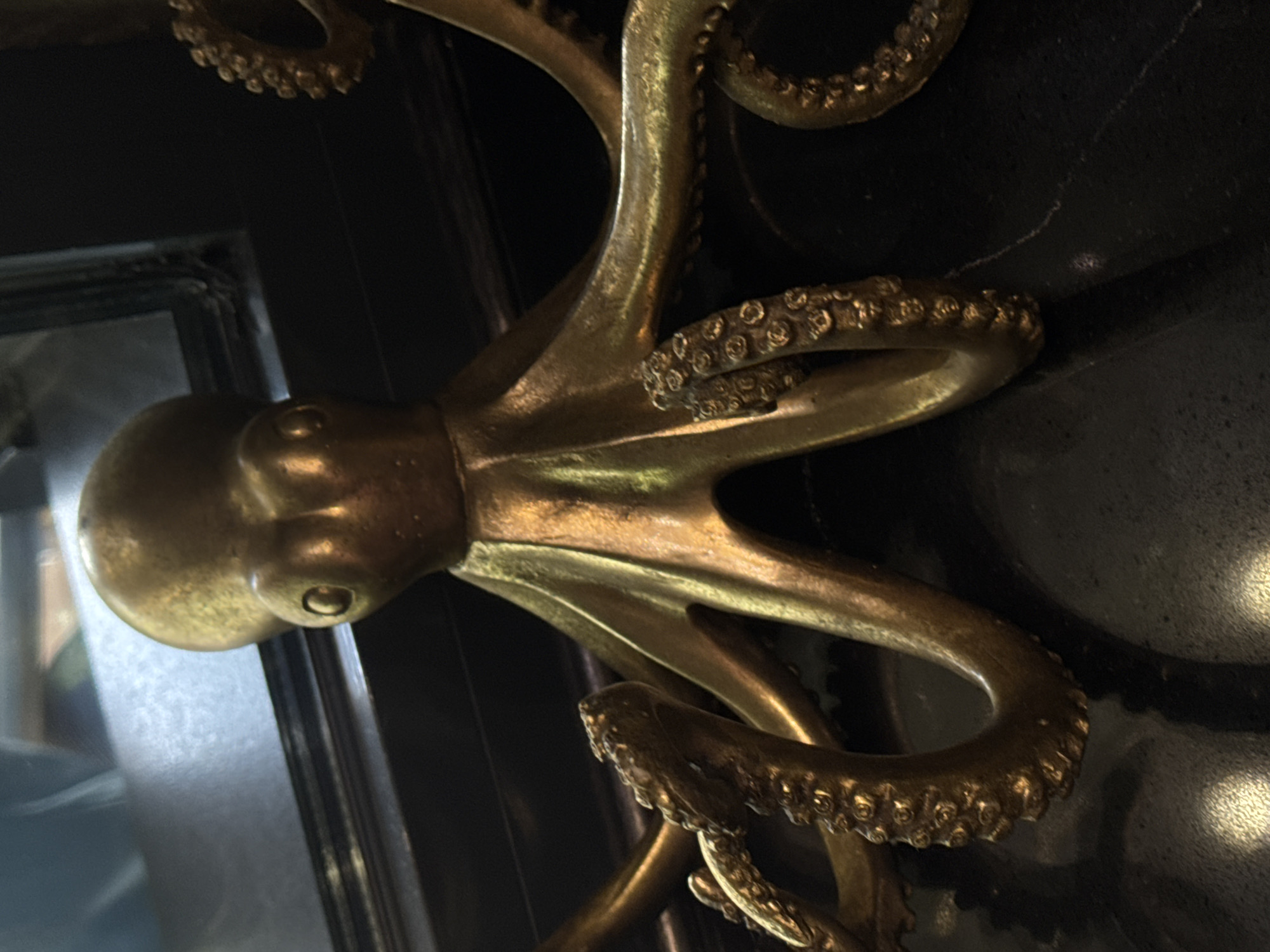 A close-up, overhead shot focuses on a golden octopus sculpture. The sculpture is positioned on a dark, reflective surface, possibly a countertop or table. The octopus has a round body with two visible eye sockets and eight tentacles. The tentacles are detailed with numerous suction cups, and some are curved in various directions, extending towards the foreground. The lighting casts strong highlights on the metallic surface of the sculpture, emphasizing its polished texture. The background is blurred but appears to show a dark frame and a reflective surface with hints of blue and green, possibly a mirror or a window. The setting is indoors, in an unknown location.