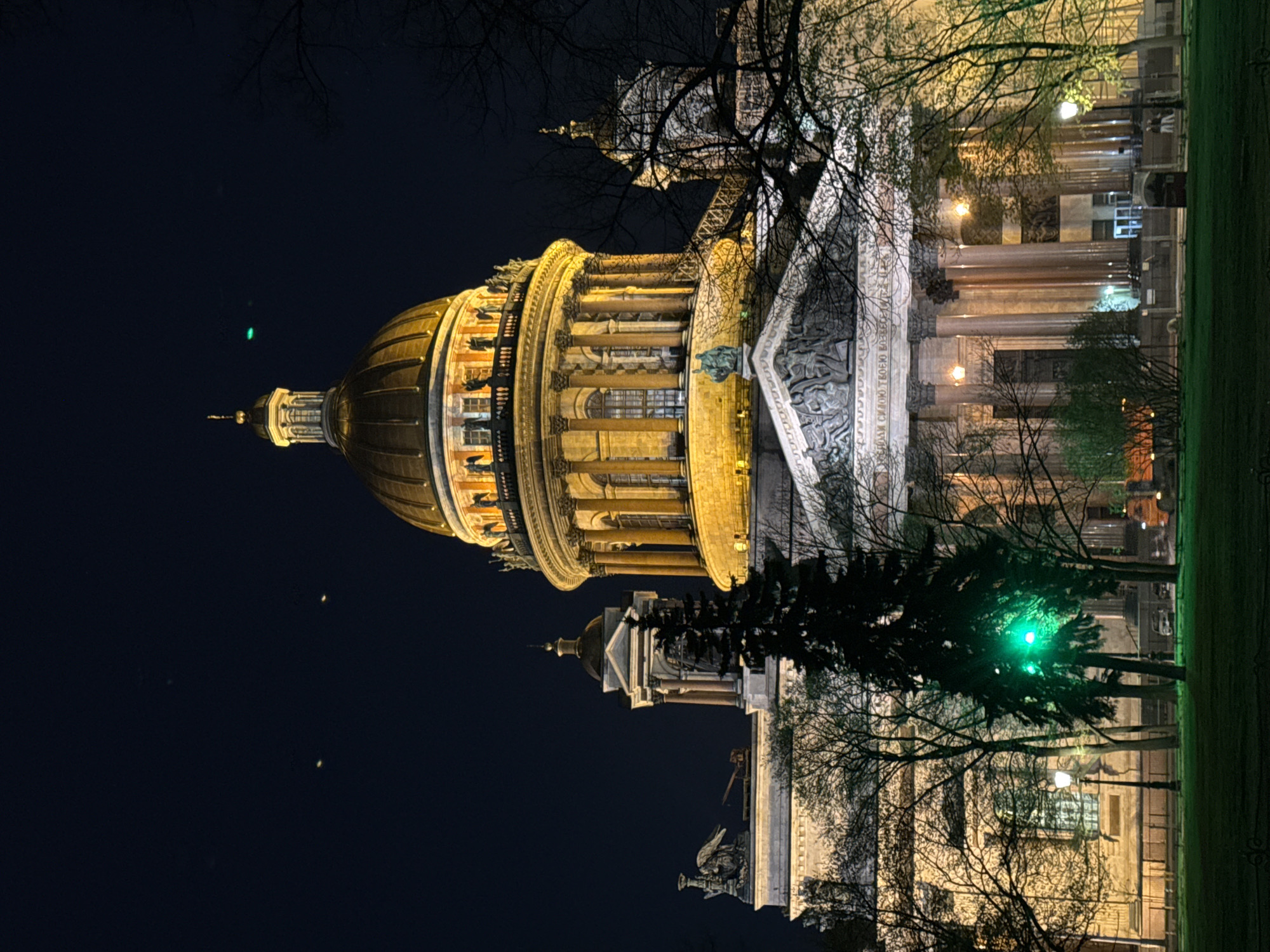 Here is a description of the image:
The image shows St. Isaac's Cathedral in Saint Petersburg, Russia, illuminated at night. The cathedral's dome is the most prominent feature, glowing with warm light. The facade of the building is also lit, showing its architectural details. Bare trees frame the building. The night sky is a deep blue, with a few faint points of light. The cathedral is seen from a low angle, emphasizing its height. A small portion of a grassy area and street lamps are visible on the right edge of the picture.