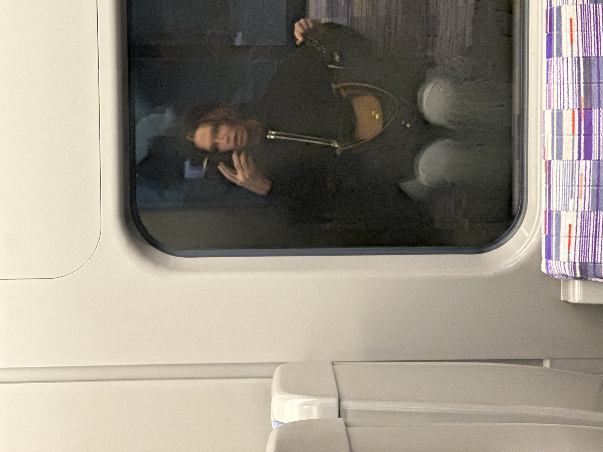 A passenger is visible through the window of a train car. The passenger, possibly male, is holding a mobile phone. The train car has a curtain with vertical stripes of white, purple, and red. The location may be London, United Kingdom.