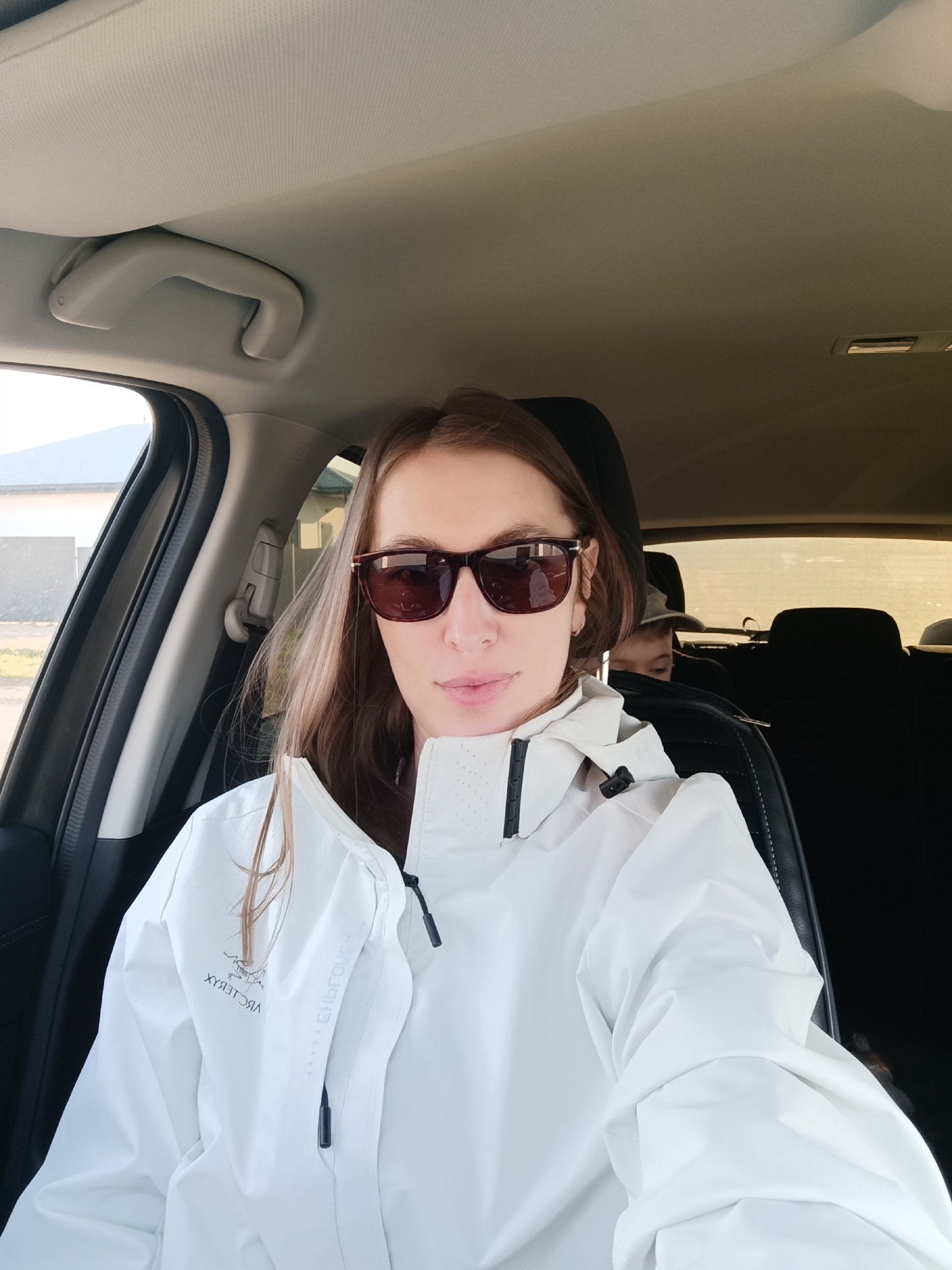 A woman in sunglasses and a white jacket is in the driver's seat of a car. A child is visible in the back seat. The interior of the car is visible, including the headliner and the passenger side grab handle. Outside the car, a building is visible. Based on the location provided, the setting is Chaikovsky, Russia.