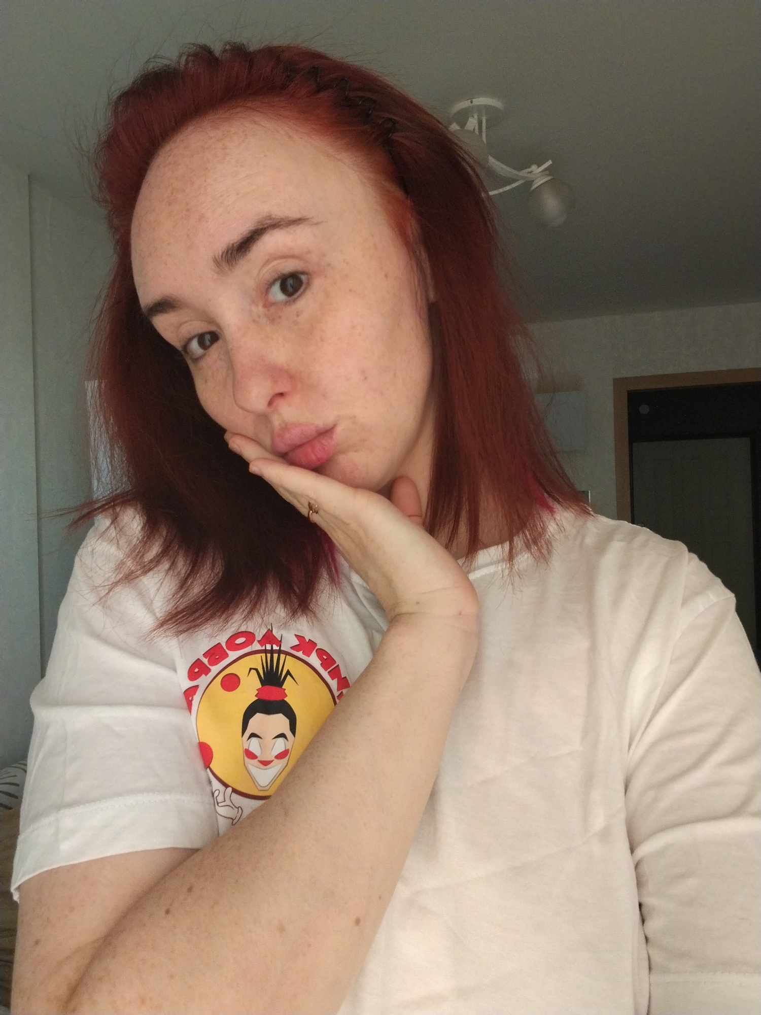 A pale-skinned woman with red hair and freckles is posing indoors. She is wearing a white t-shirt with a colorful cartoon graphic on the front. Her left hand is touching her face, and she is puckering her lips. The background includes a white wall, a ceiling with a light fixture, and a door. There is no information provided that would lead to determining the location to be Izhevsk, Russia.