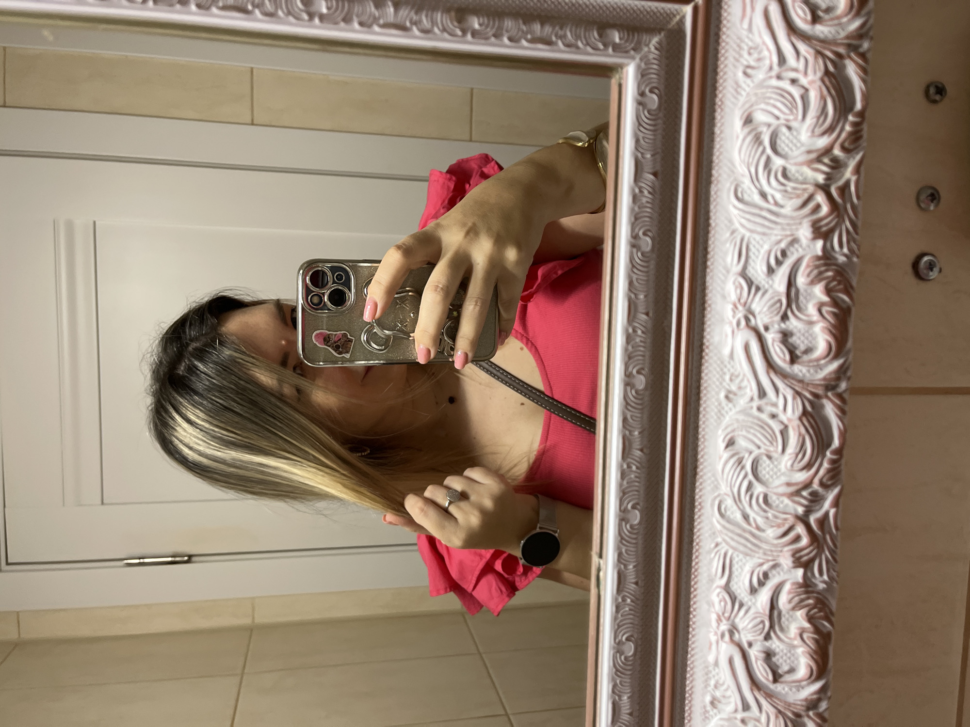 A female individual is captured in a mirror selfie in a room located in Sochi, Russia. The woman, shown from the chest up, has dark hair with prominent blonde highlights, particularly framing her face. Her face is partially obscured by the metallic, multi-lens iPhone she holds in her right hand, which also features a pink-hatted cat sticker and another partially visible cartoon character sticker. Her visible eye appears to be looking towards the phone screen.
She wears a vibrant fuchsia-pink top; the left shoulder has ruffled, layered fabric, while the main body appears ribbed. On her left hand, a silver-toned ring with a clear, circular stone adorns her ring finger, and a silver mesh-band smartwatch with a black, inactive screen is on her wrist. This hand is positioned near her head, gently touching strands of her hair. Her nails are painted a light pink. On her right wrist, a gold-toned bracelet with a white oval accent is visible. A thin, dark necklace is visible around her neck.
The background, reflected in the ornate mirror, reveals an interior space. To the left, light beige ceramic tiles with vertical grout lines are visible. To the right, a white paneled interior door with a visible silver hinge is present. The mirror's frame is highly decorative, featuring intricate carved scrollwork, floral motifs, and organic patterns. The frame is painted an off-white or light cream color, with subtle pinkish undertones in its recesses, and is bordered by a thinner inner trim of a brown or bronze hue. Below the mirror frame, a light-colored surface with three small, circular, silver-colored elements (possibly screw caps or decorative studs) is visible.