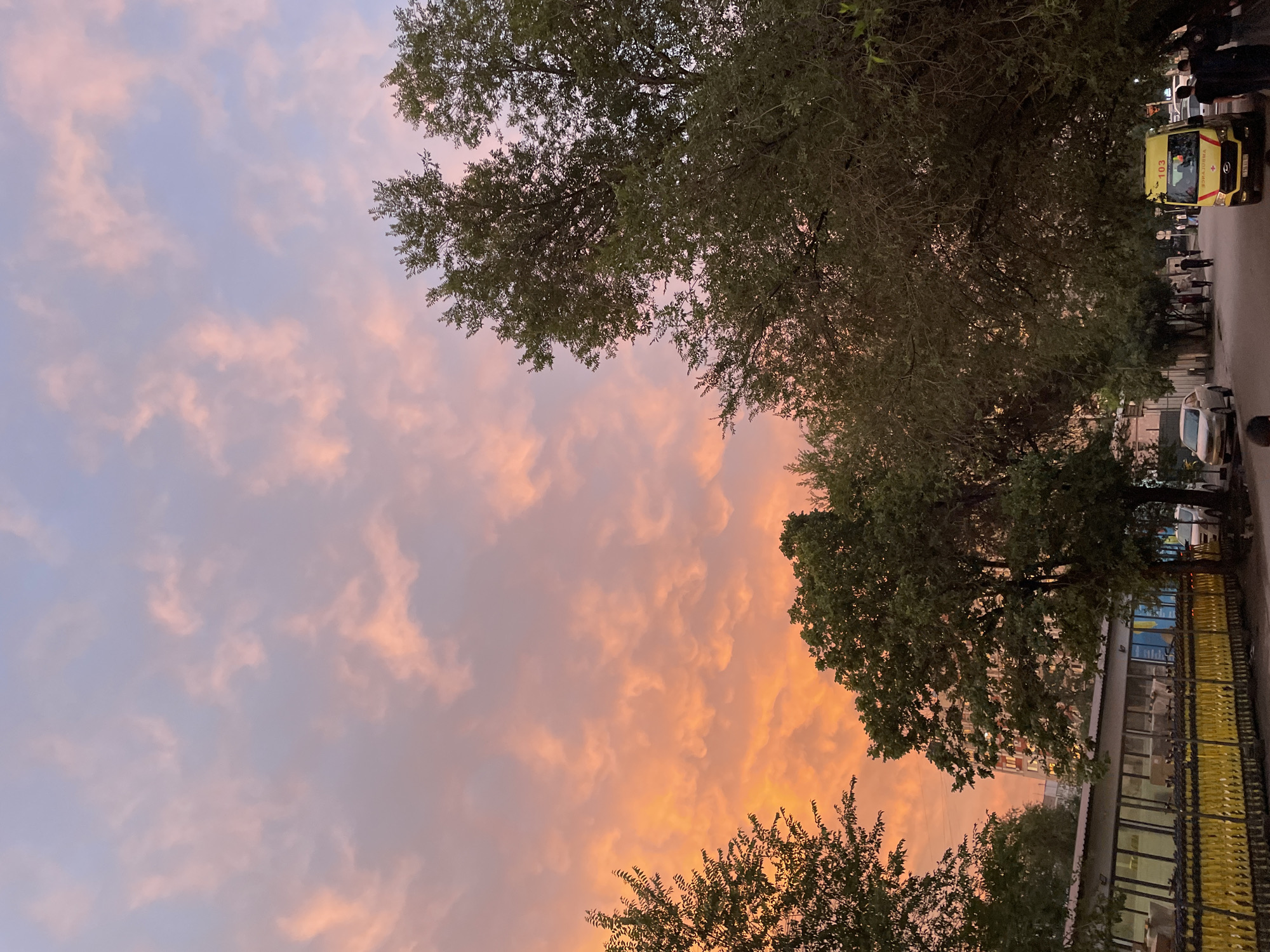 This image captures a street scene in Almaty, Kazakhstan, during sunset. The sky is filled with clouds tinged with orange and pink hues, contrasting with the soft blue of the upper atmosphere. In the foreground, trees with dense green foliage line the street, partially obscuring buildings and the sky.
On the right side of the street, a yellow ambulance with the number "103" prominently displayed is parked. Several individuals, some appearing to be medical personnel in dark uniforms, are standing near the ambulance. Further down the street, other pedestrians can be seen walking.
To the left, a row of yellow carts or barriers is visible along a sidewalk or building facade. The street itself is paved, and a few cars are parked or driving on it. The background reveals apartment buildings, suggesting an urban environment. The overall impression is one of a quiet street moment captured as day transitions into evening.