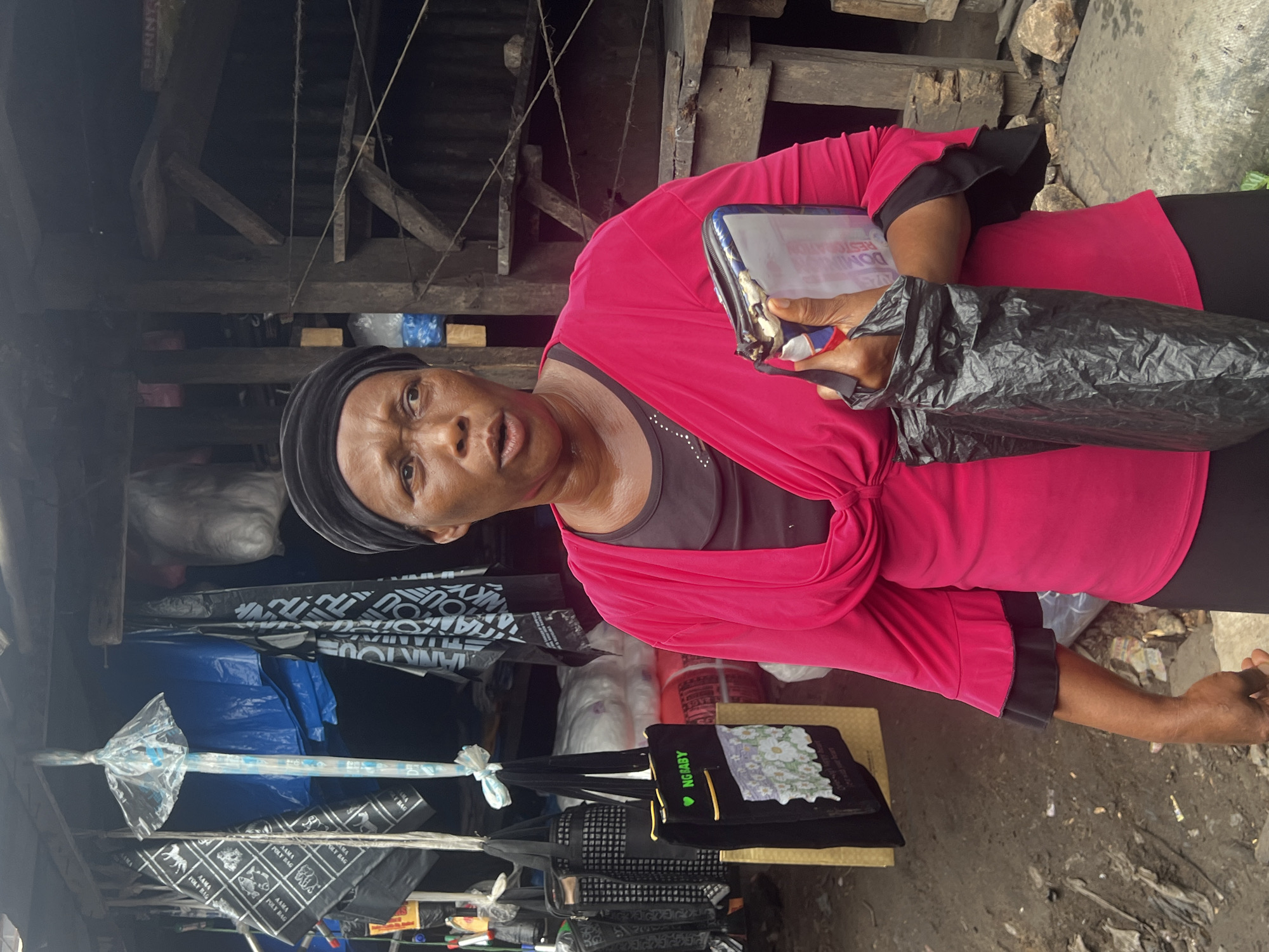 In Nkpor, Nigeria, a woman wearing a black headscarf and a bright pink top with flared black cuffs stands outdoors. She is holding a crumpled black plastic bag in her right hand and a small zippered pouch in her left hand, which appears to contain documents or money. The background shows a rustic market stall with wooden structures and various items for sale hanging from the ceiling, including blue tarps, black bags with white text, and a black tote bag with a floral design and the text "NG BABY." The ground is muddy and littered with debris, suggesting a potentially unpaved or unkempt area common in some market environments.