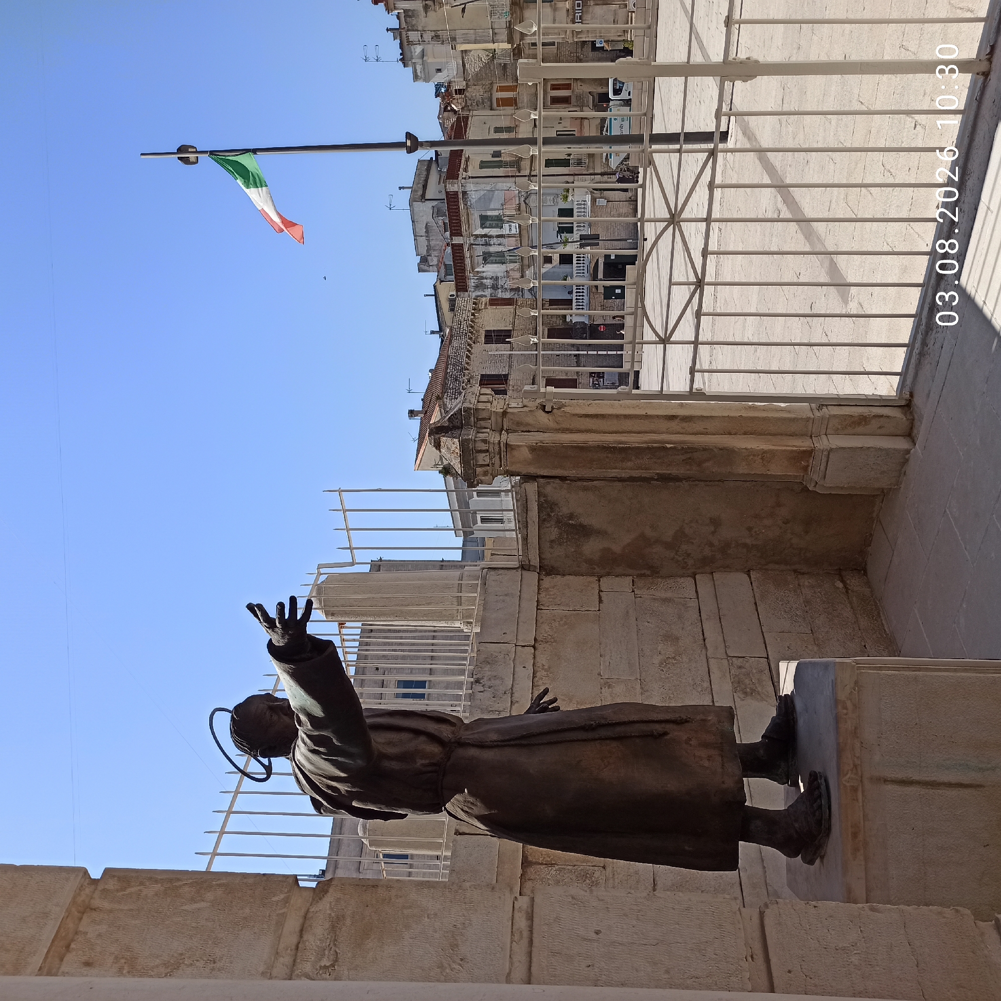 The image depicts a bronze statue of a robed figure with a halo, positioned outdoors in a European city square. The statue is placed on a stone pedestal, with its right arm extended forward and its left arm bent at the elbow. The background shows a bright blue sky, a white metal fence, and several buildings with traditional architecture, including tiled roofs and balconies. An Italian flag is visible flying from a flagpole in the distance. The foreground shows a paved surface and a timestamp "03.08.2026 10:30" in white lettering. The statue is likely located in Ruvo di Puglia, Italy, as it is a common subject of images from that city. The scene appears to be a sunny day.