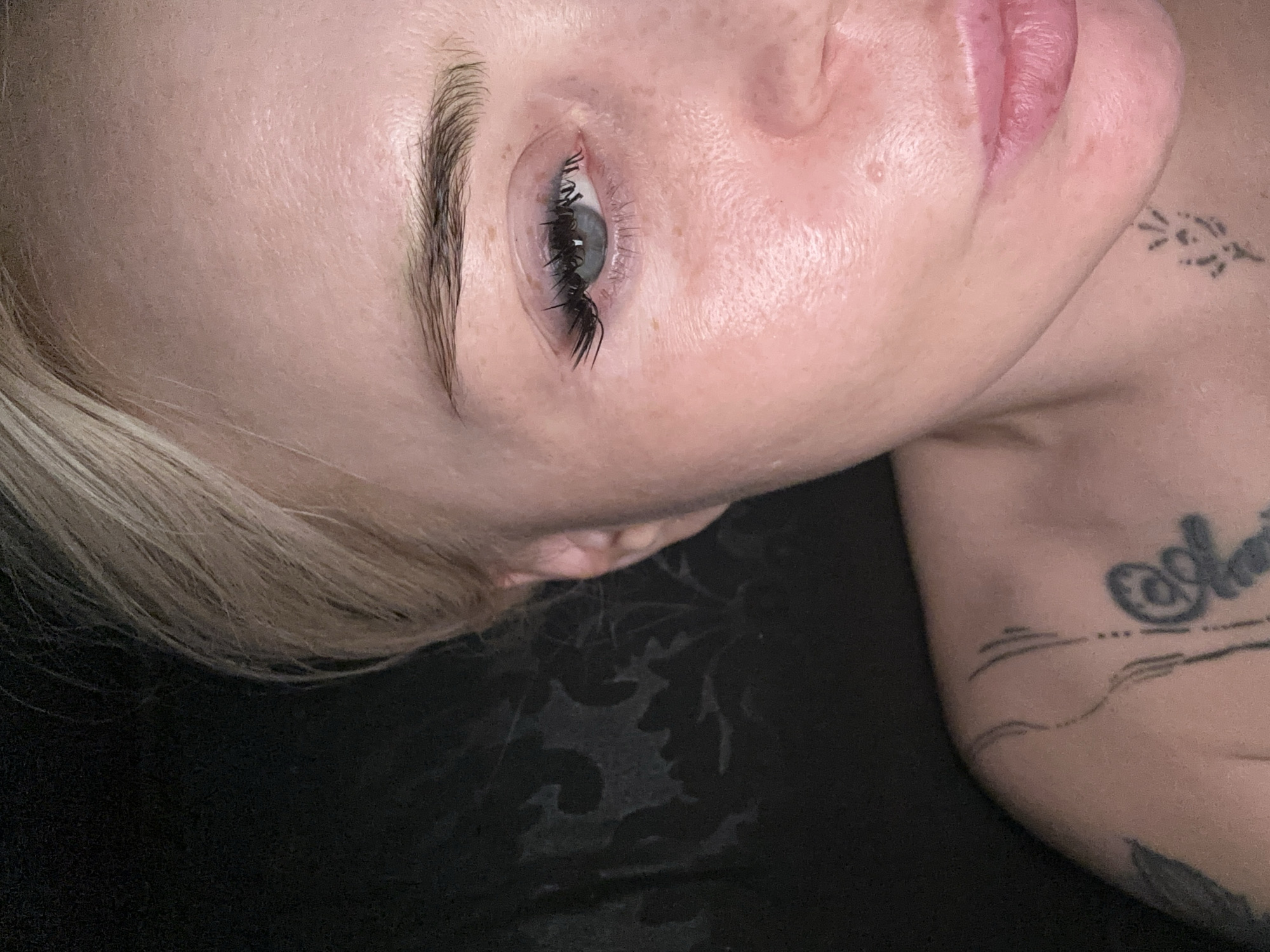 A close-up, rotated shot captures a portion of a person's upper face and shoulder against a dark background. The subject has fair skin and light-colored hair, visible on the left side of the frame. The central focus is on a light blue eye, which is open and looking slightly upward. The eyelashes are prominent, dark, long, and curled, suggesting they have been enhanced or treated. Above the eye, a well-defined, dark eyebrow is visible.
A portion of the person's lips, appearing naturally pink and full, is visible on the upper right side of the frame. On the person's right shoulder and upper arm, several dark tattoos are present. These include fine lines, abstract patterns, and what appears to be stylized text, though the specific words are not clearly legible.
The background is a dark, possibly black, fabric with a subtle, dark floral or damask pattern. The overall scene suggests an indoor setting, with the person likely lying down or reclining. No specific activity is depicted beyond a still pose. The lighting is soft and even, highlighting the skin texture and features.
