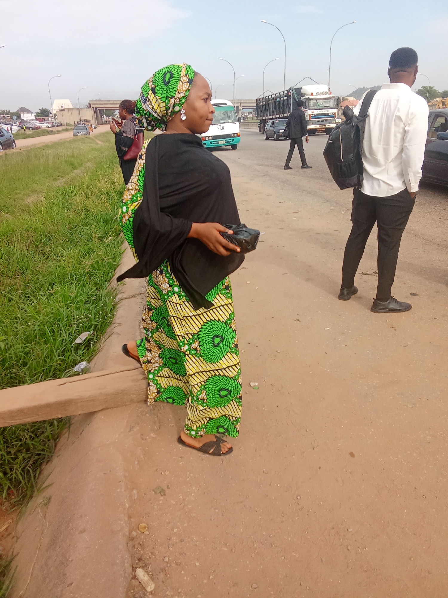 The image depicts a street scene in Kagini, Nigeria, on an overcast day. A woman in a black headscarf and a green, yellow, and white patterned dress stands on a dirt path next to a grassy verge. She is holding a black clutch bag. To her right, a man in a white long-sleeved shirt and black trousers walks away from the camera, carrying a black backpack over his right shoulder. In the background, a white bus with "NAN" on the front is visible, along with several trucks and cars on a road. A pedestrian overpass is also present in the background, suggesting an urban or semi-urban environment. The overall scene is a snapshot of everyday life on a Nigerian street, with a focus on the pedestrians present.