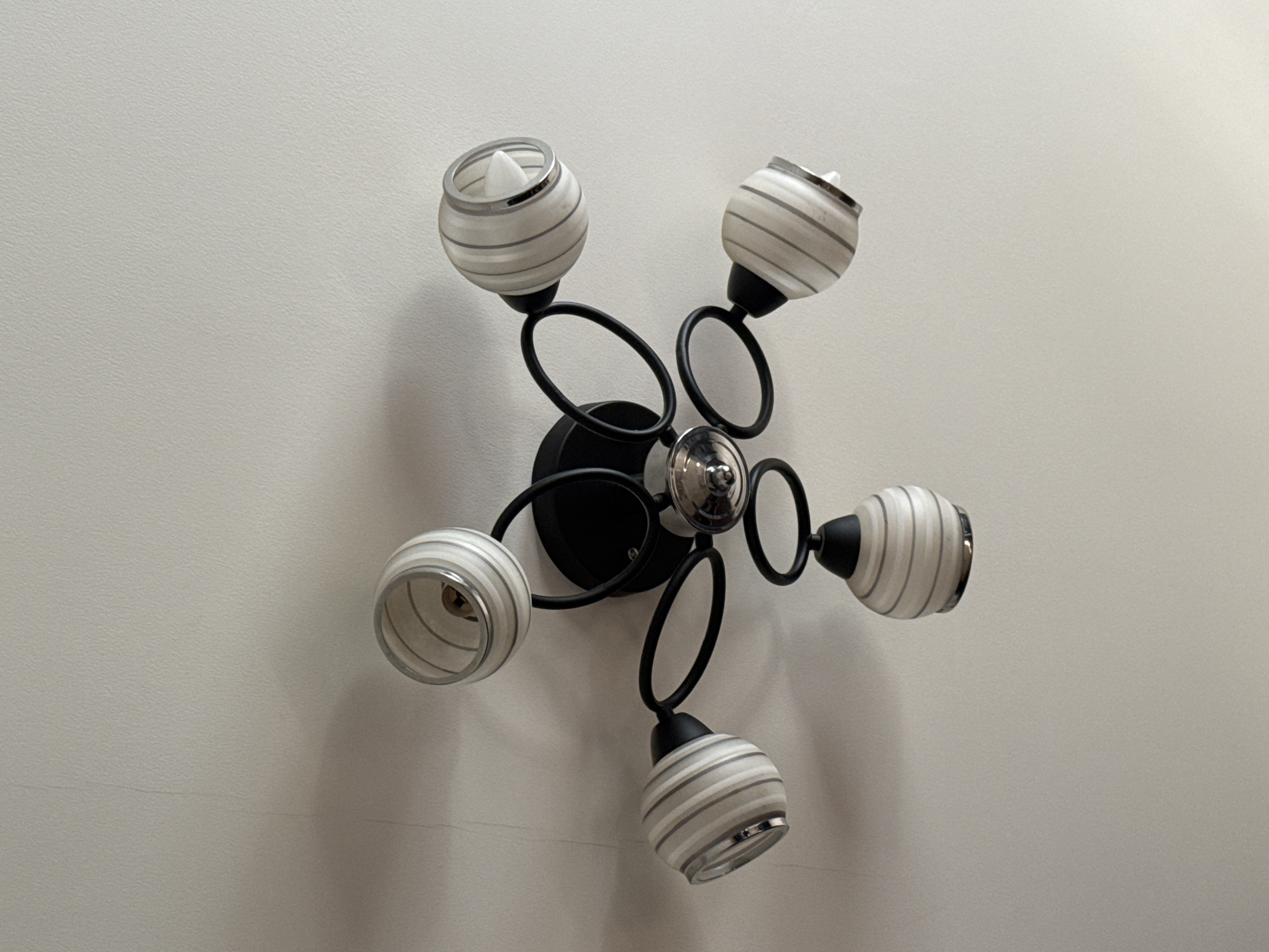 The image shows a modern ceiling light fixture with five arms extending from a central black base. Each arm is adorned with a circular metal loop and ends in a light socket covered by a cylindrical glass shade. The shades have a white base with thin, horizontal gray stripes. The ceiling is painted white and shows a faint crack near the top left corner, with a subtle shadow cast by the light fixture. The scene appears to be indoors, likely in a residential setting. There are no people or animals visible in the image.